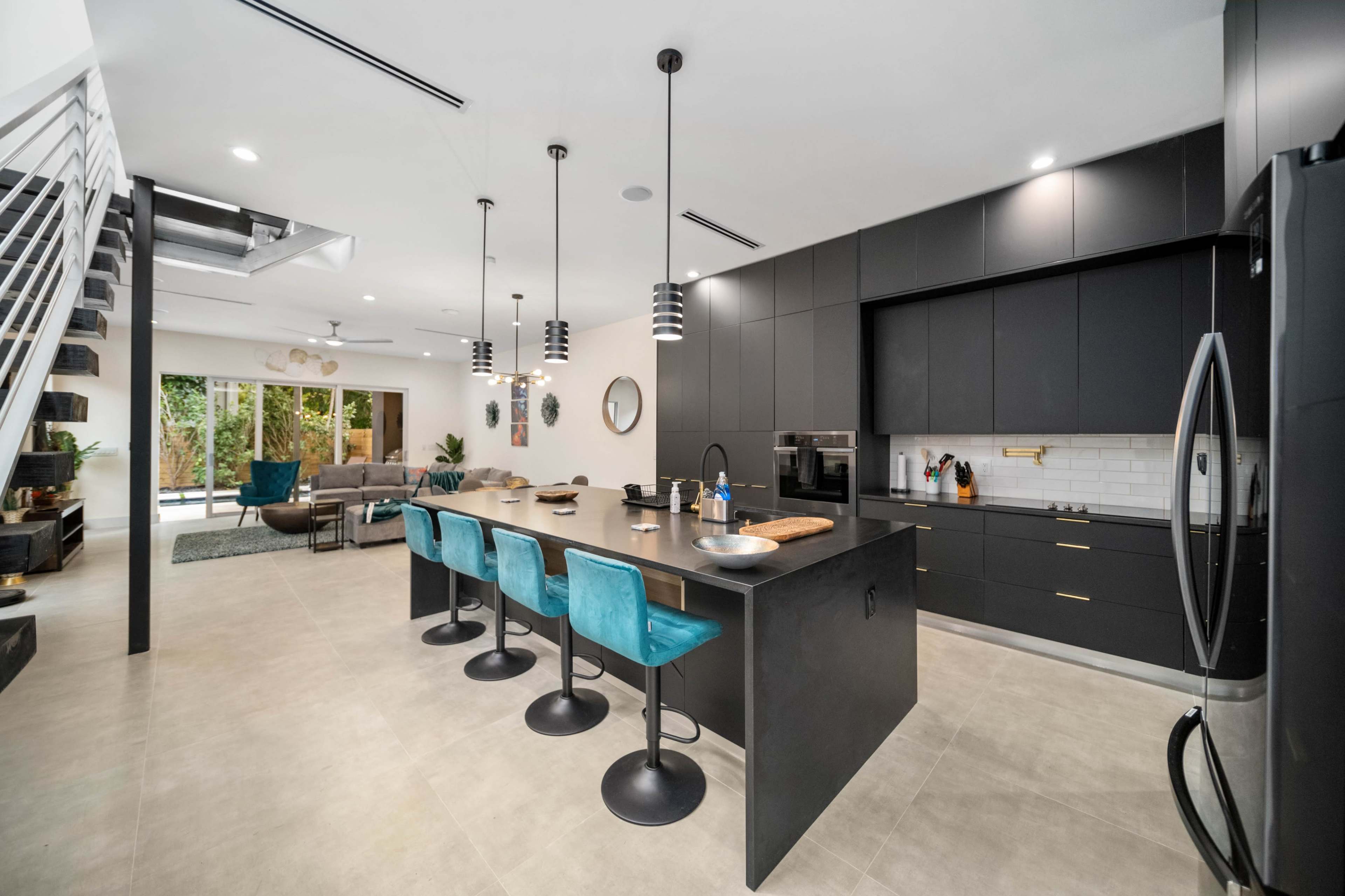 A modern kitchen features a large black island with blue bar stools, stainless steel appliances, and opens to a living area with large windows.