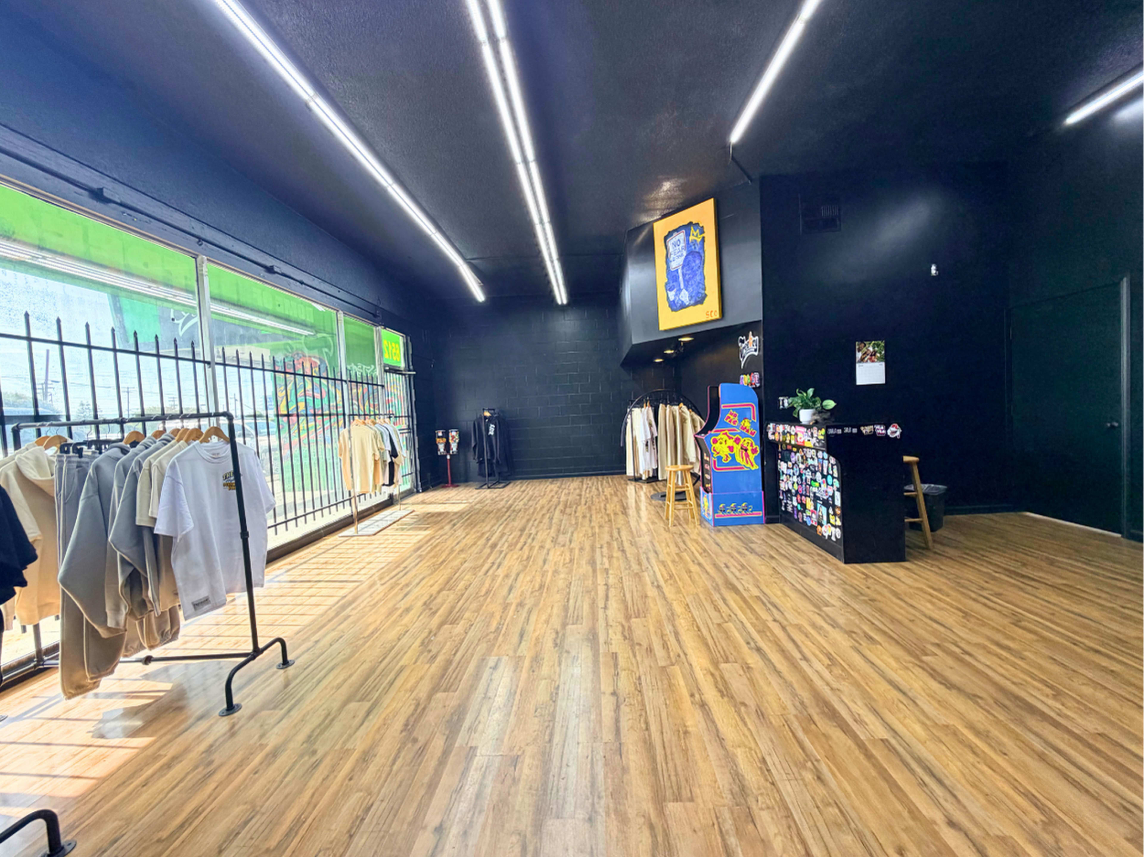 The image depicts an empty clothing retail space with wooden flooring, black walls, and various clothing items displayed on racks along one side.