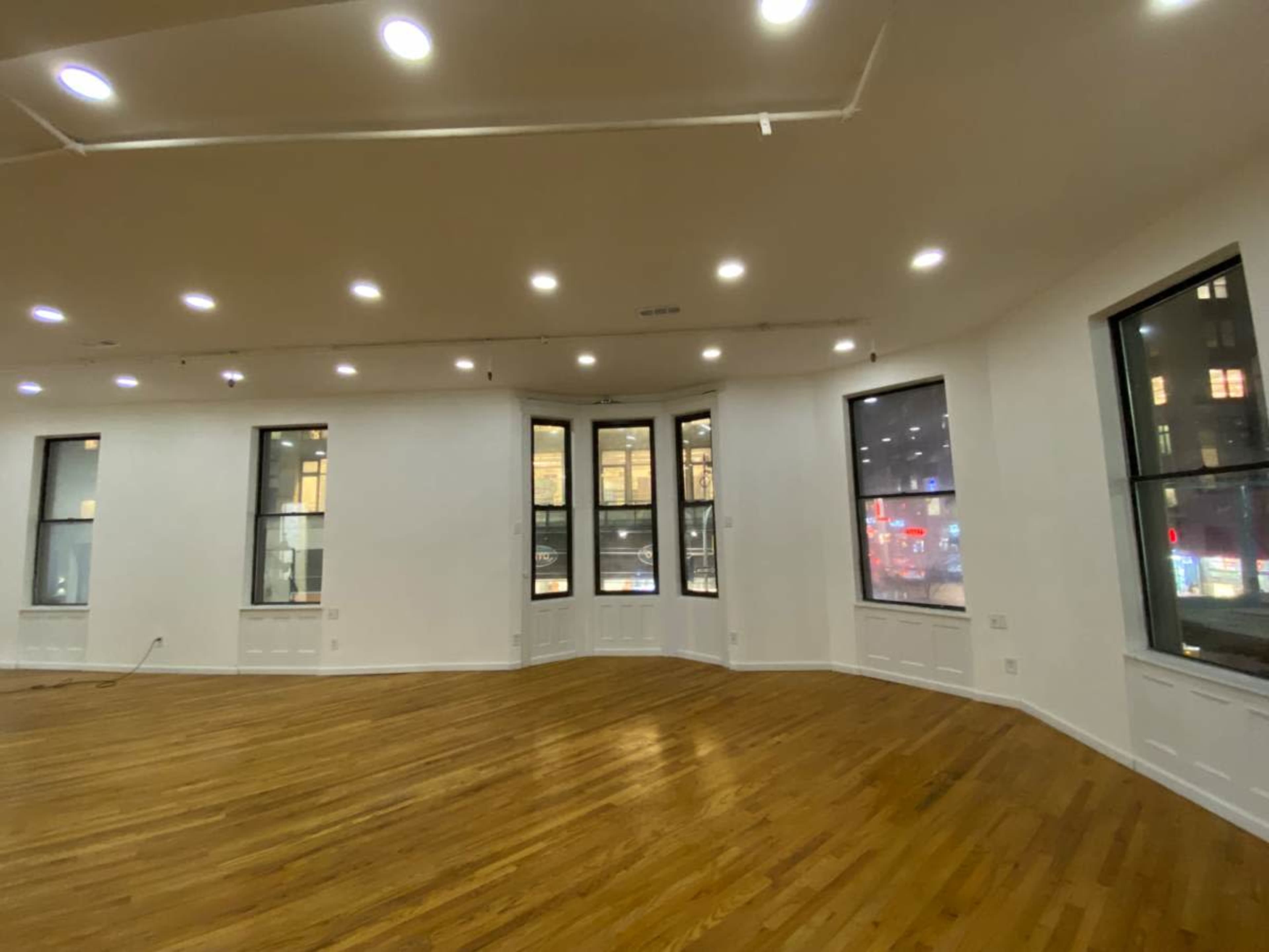 The image shows an empty room with hardwood floors, featuring large windows and bright ceiling lights.