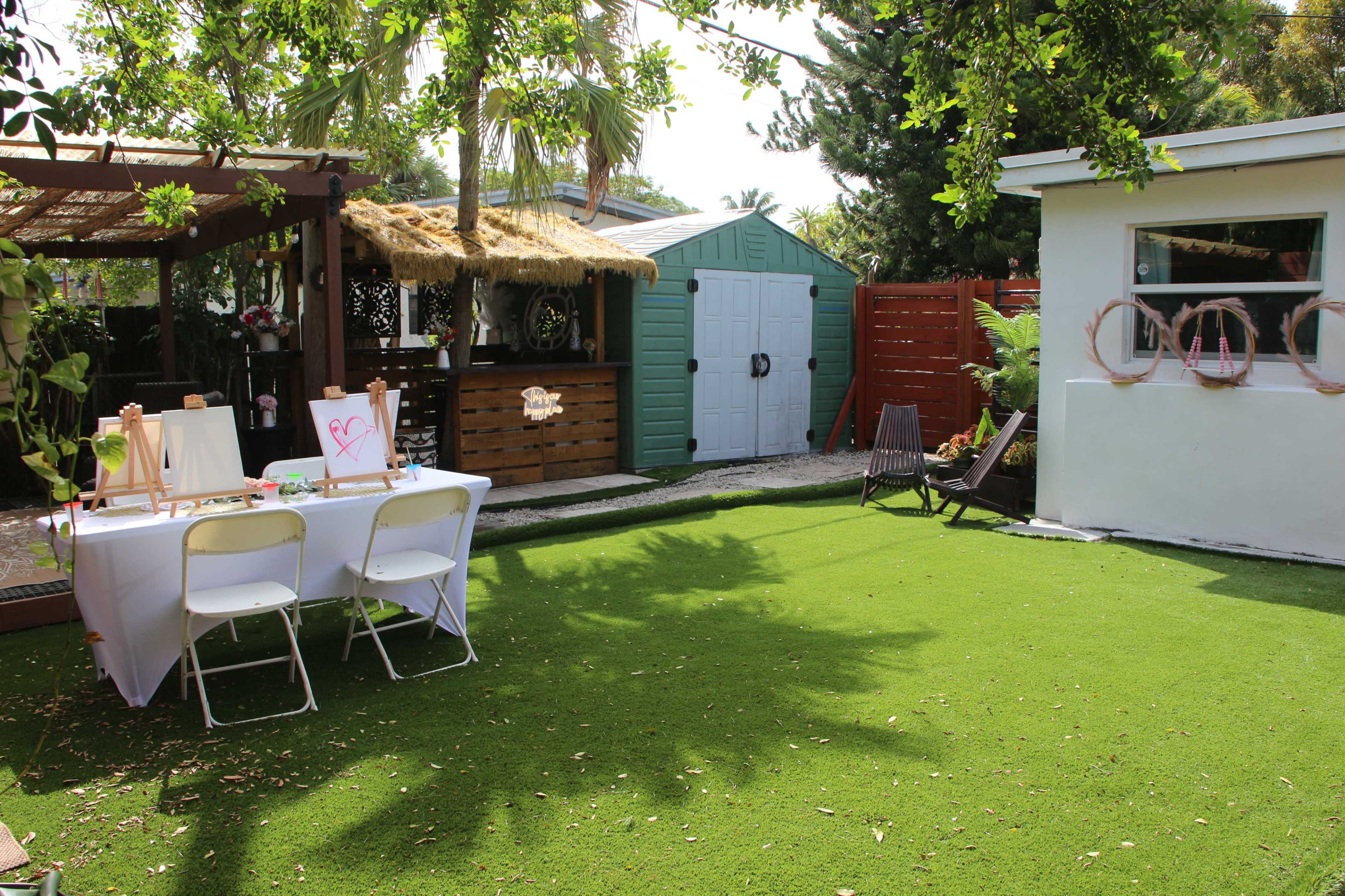The scene features a green outdoor area with a table set up covered by a white cloth, surrounded by tropical plants, a small shed, and a relaxation chair.