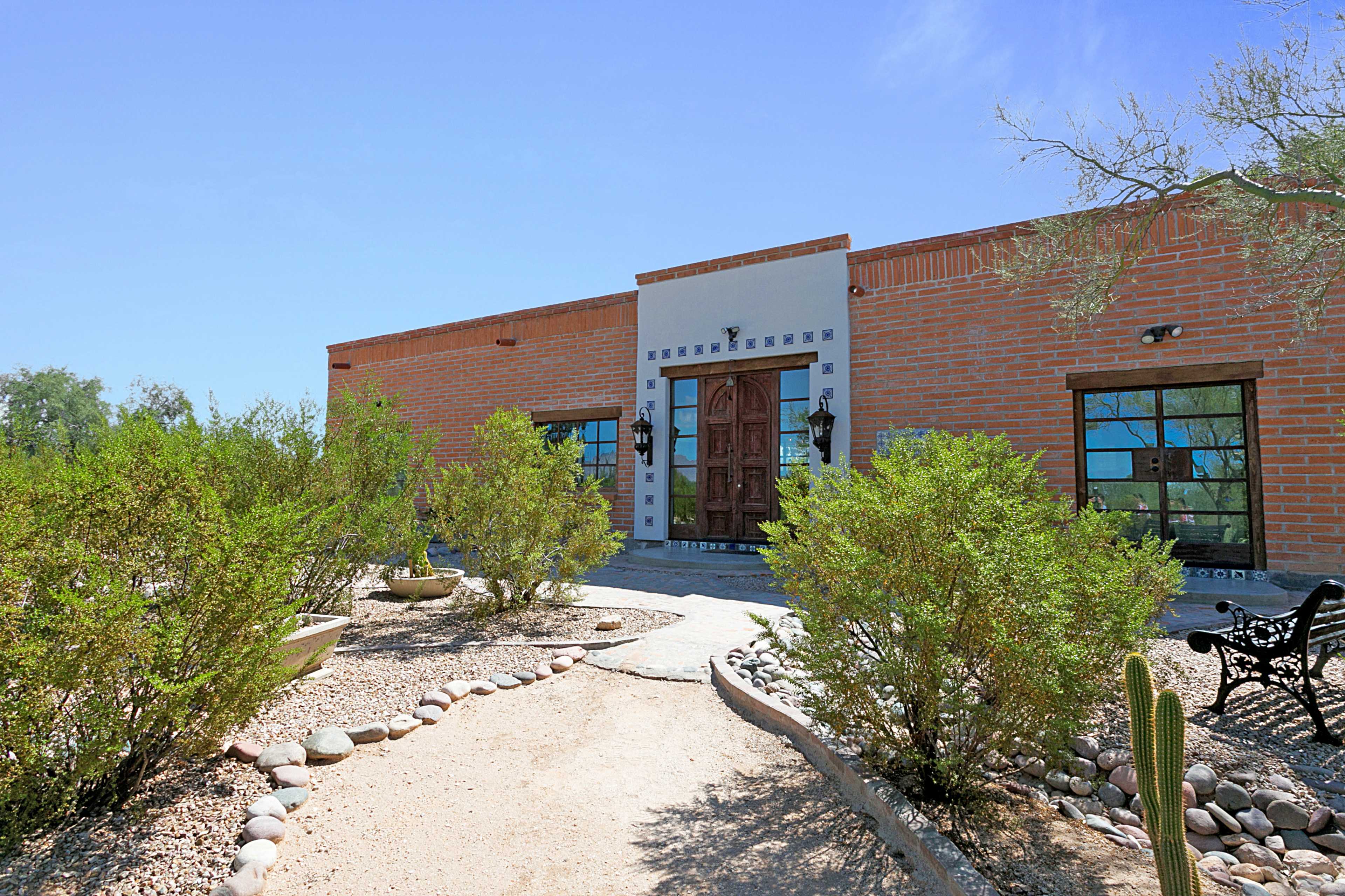 A single-story adobe-style house with a wooden front door is surrounded by desert landscaping featuring shrubs, stones, and a pathway.