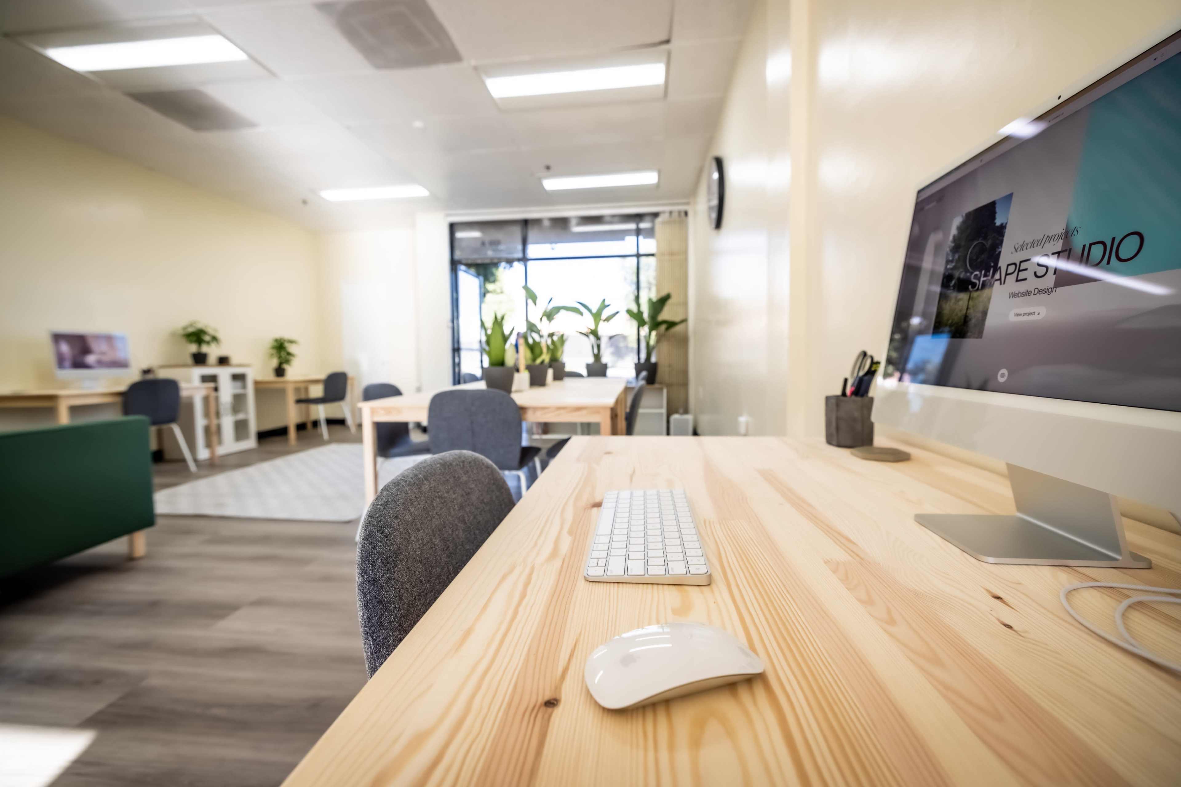 A modern office space features a wooden desk with a computer, keyboard, and mouse, surrounded by plants and furniture.