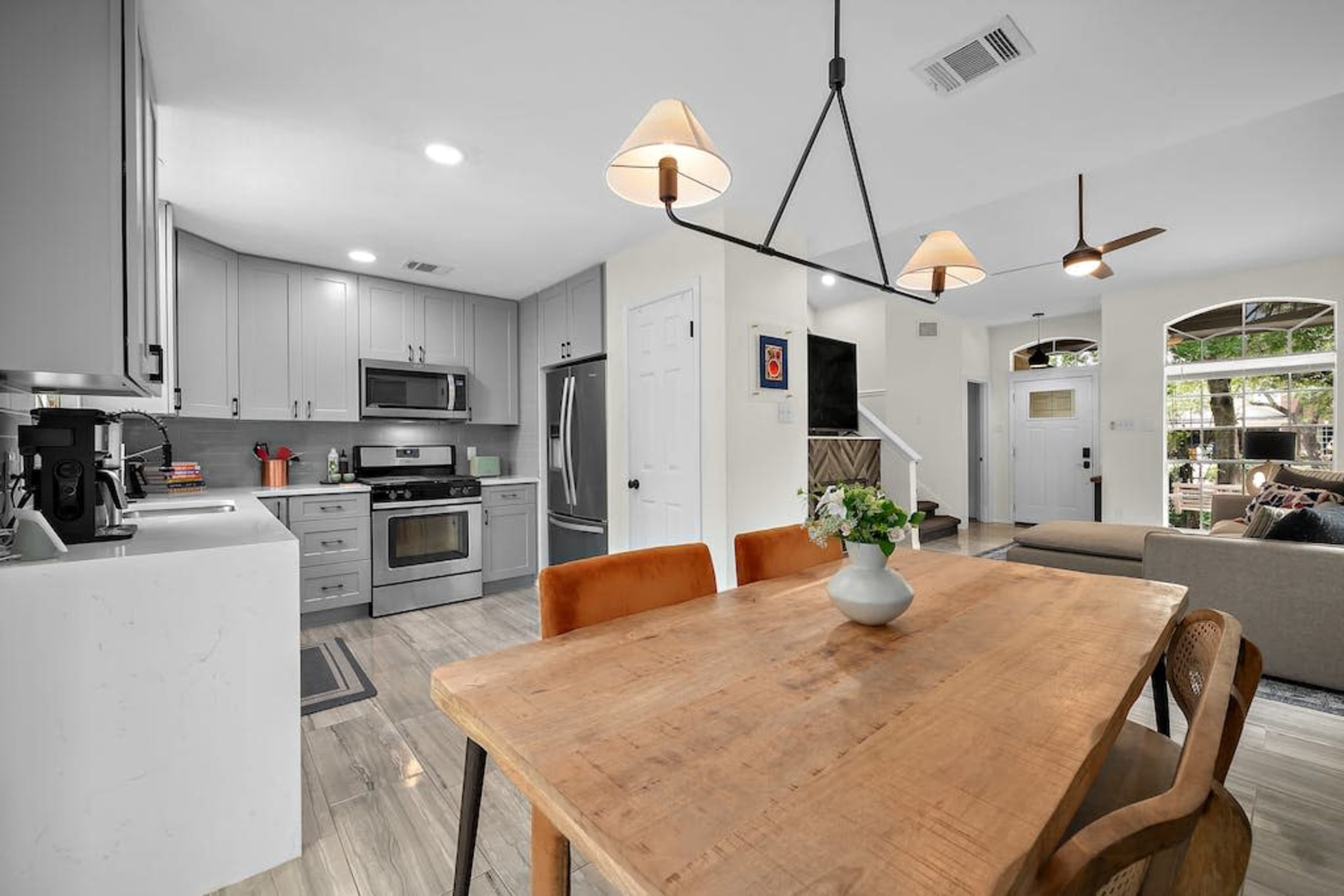 The image shows a modern kitchen and dining area with a wooden table, gray cabinetry, stainless steel appliances, and a view of a living room through an opening featuring large windows.