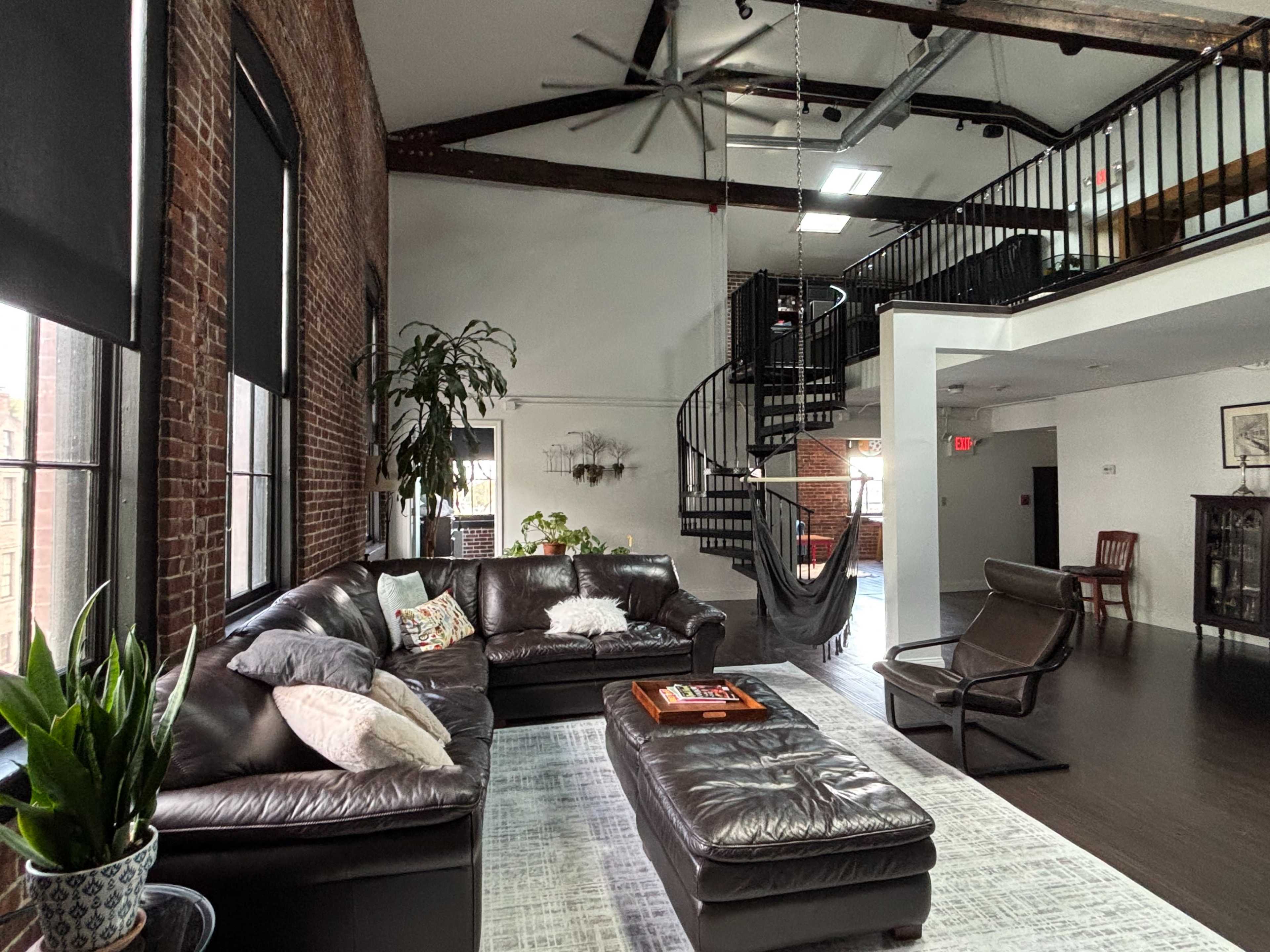 The image shows a spacious living area with a large leather sectional sofa, a spiral staircase, and hardwood floors, highlighted by natural light from large windows.