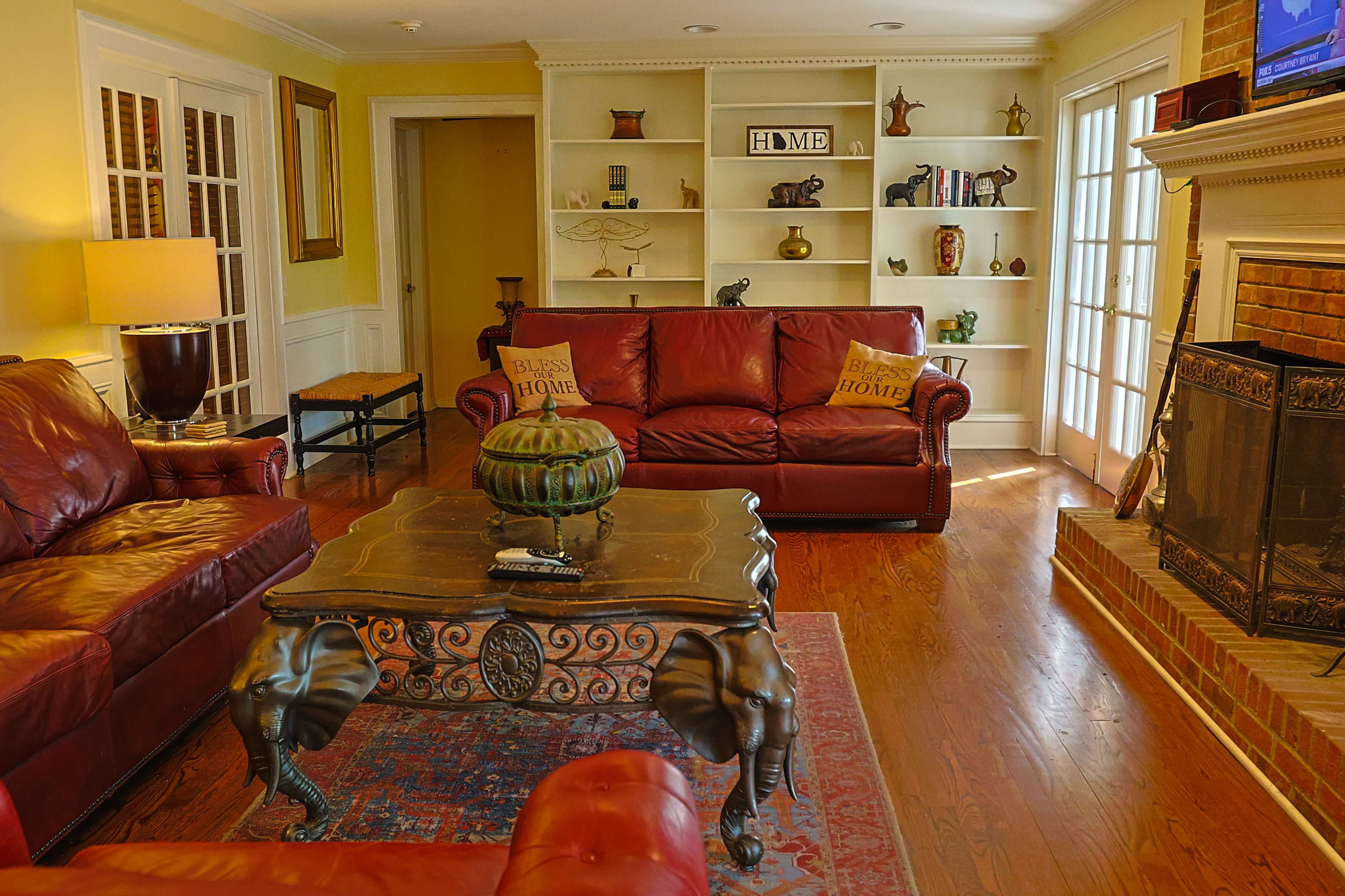 The image shows a cozy living room featuring two red leather sofas, a wooden coffee table with an elephant design, and a fireplace surrounded by built-in shelves displaying decorative items.
