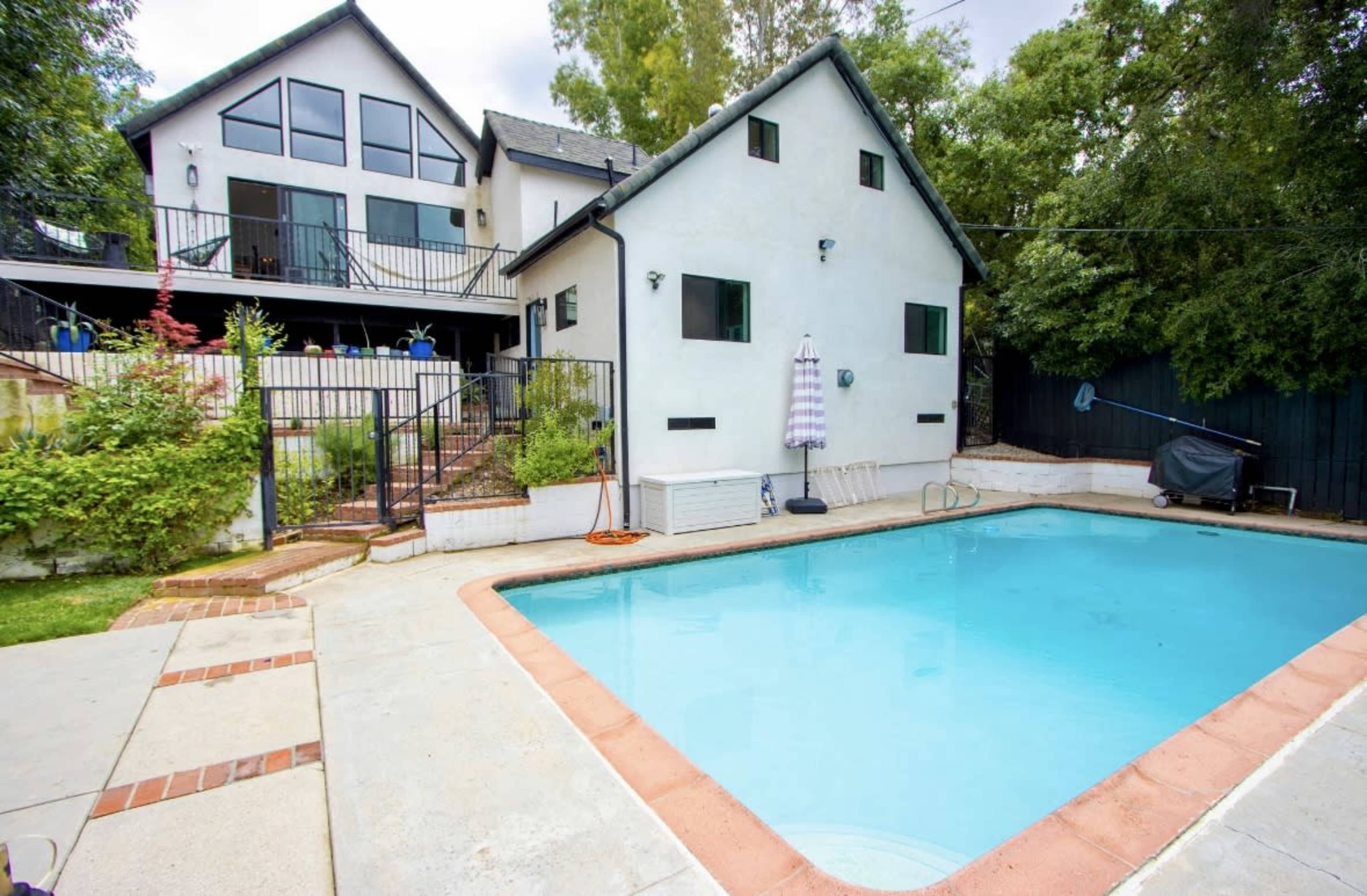 A rectangular swimming pool is situated in front of a two-story house with a landscaped yard and a staircase leading to a deck on the upper level.
