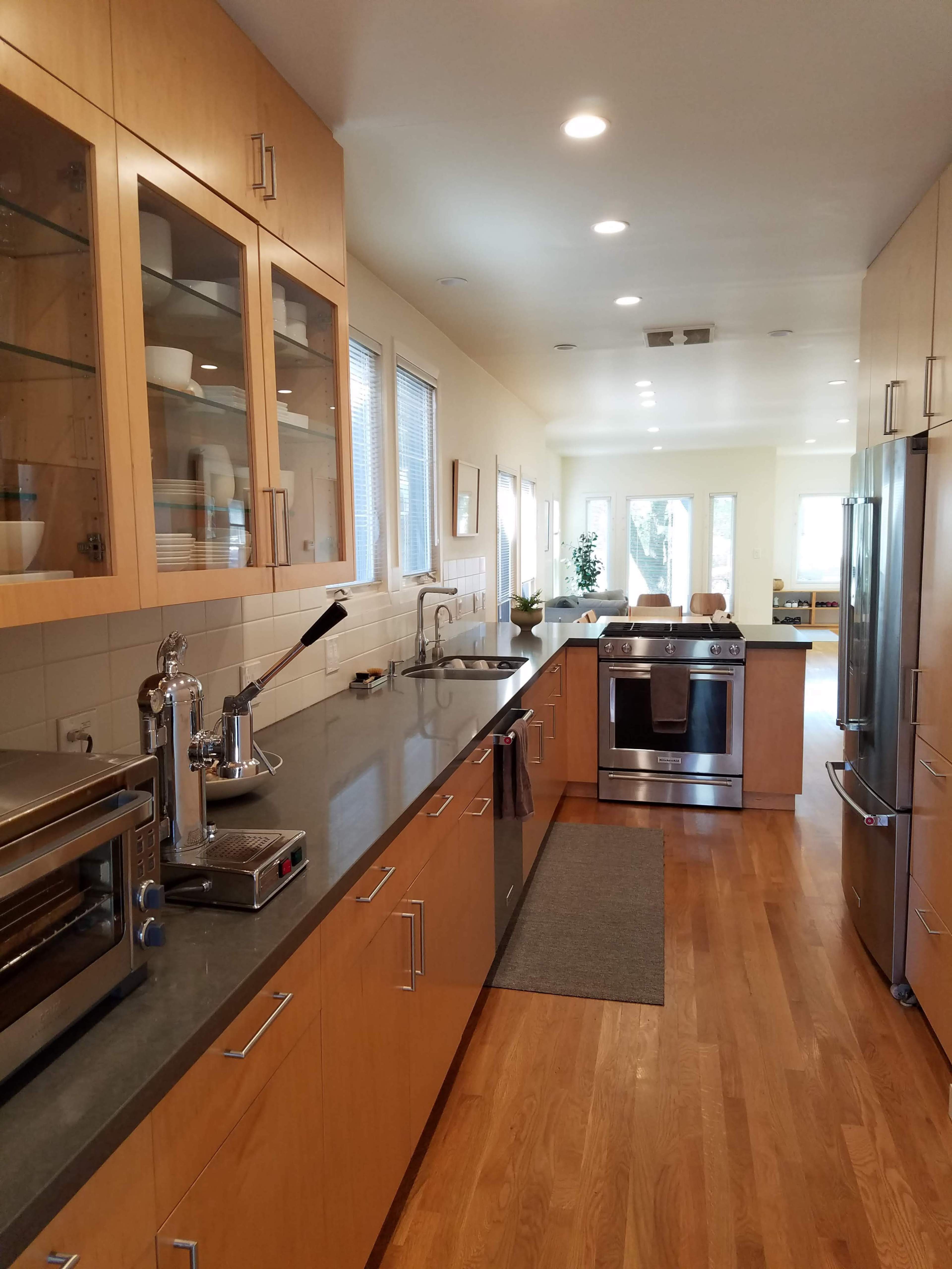 A modern kitchen features wooden cabinetry, a gray countertop, and stainless steel appliances, with an open view to a living area at the far end.