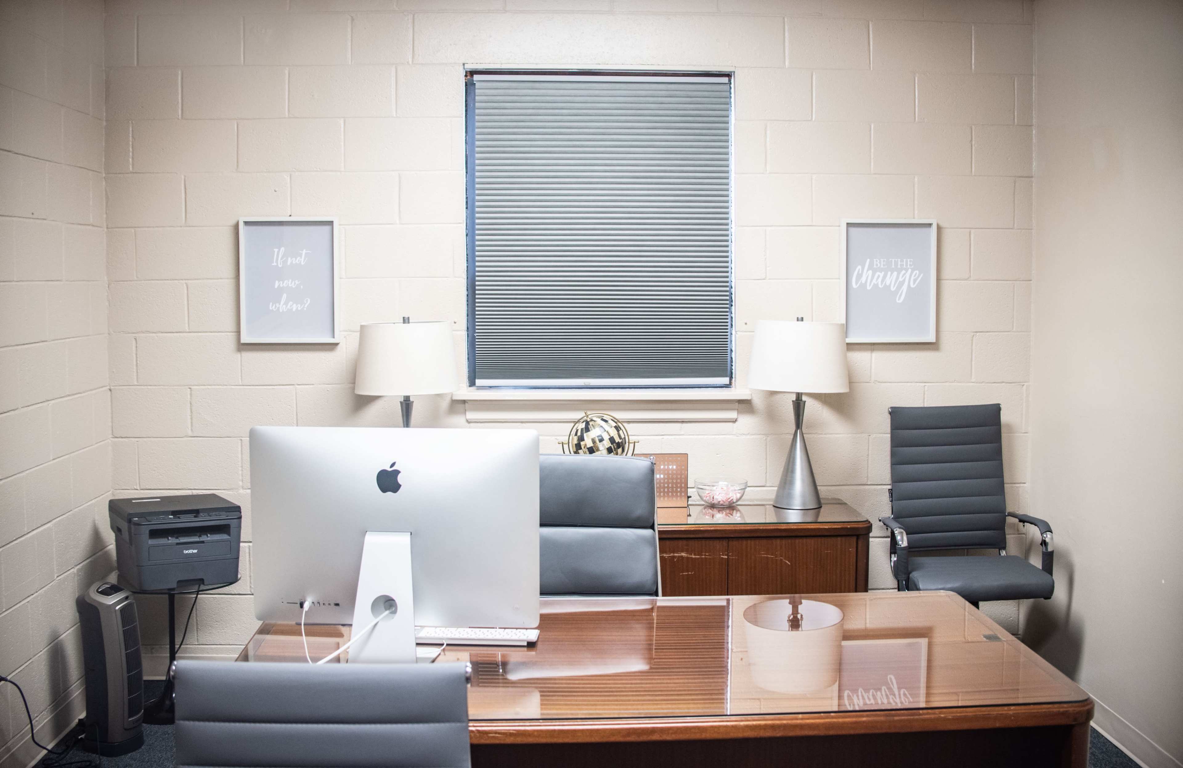 A modern office interior with a wooden desk, an iMac computer, two chairs, lamps, and motivational framed prints on the walls.