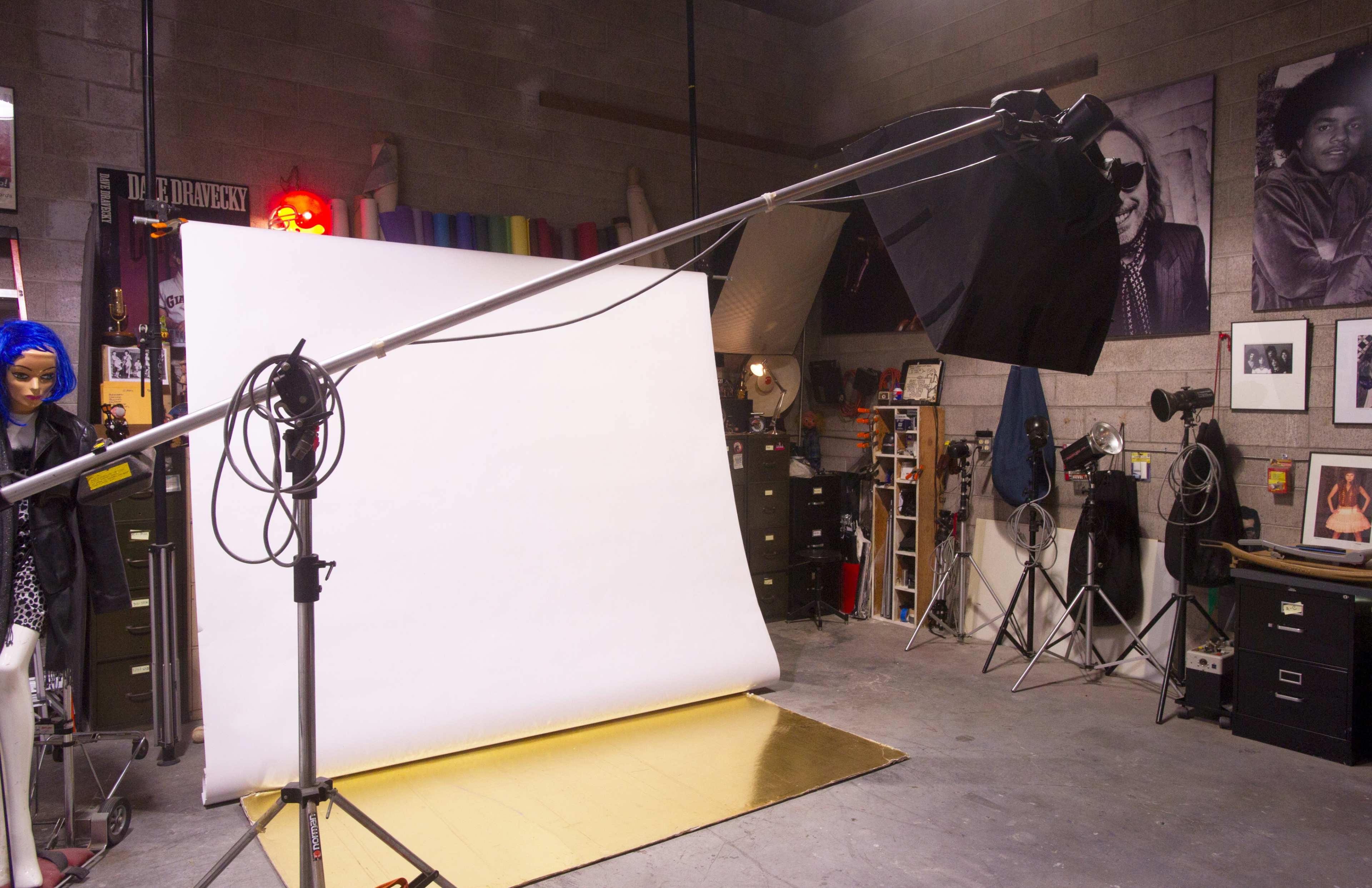 A photography studio features a white backdrop on a stand, a golden floor, and various lighting equipment, with a mannequin positioned nearby.