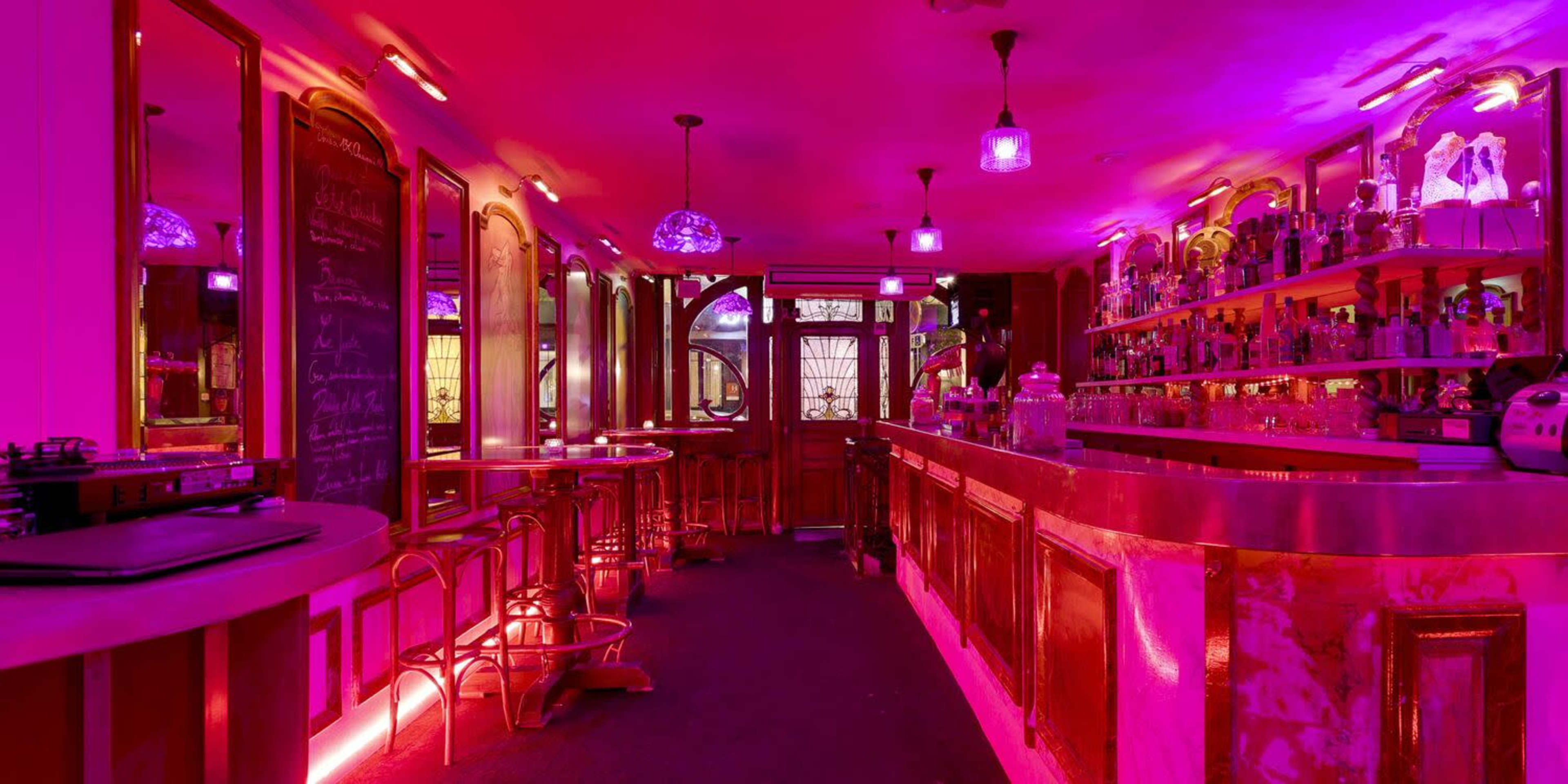 A bar with a pink-lit interior, featuring a long counter and shelves lined with bottles.