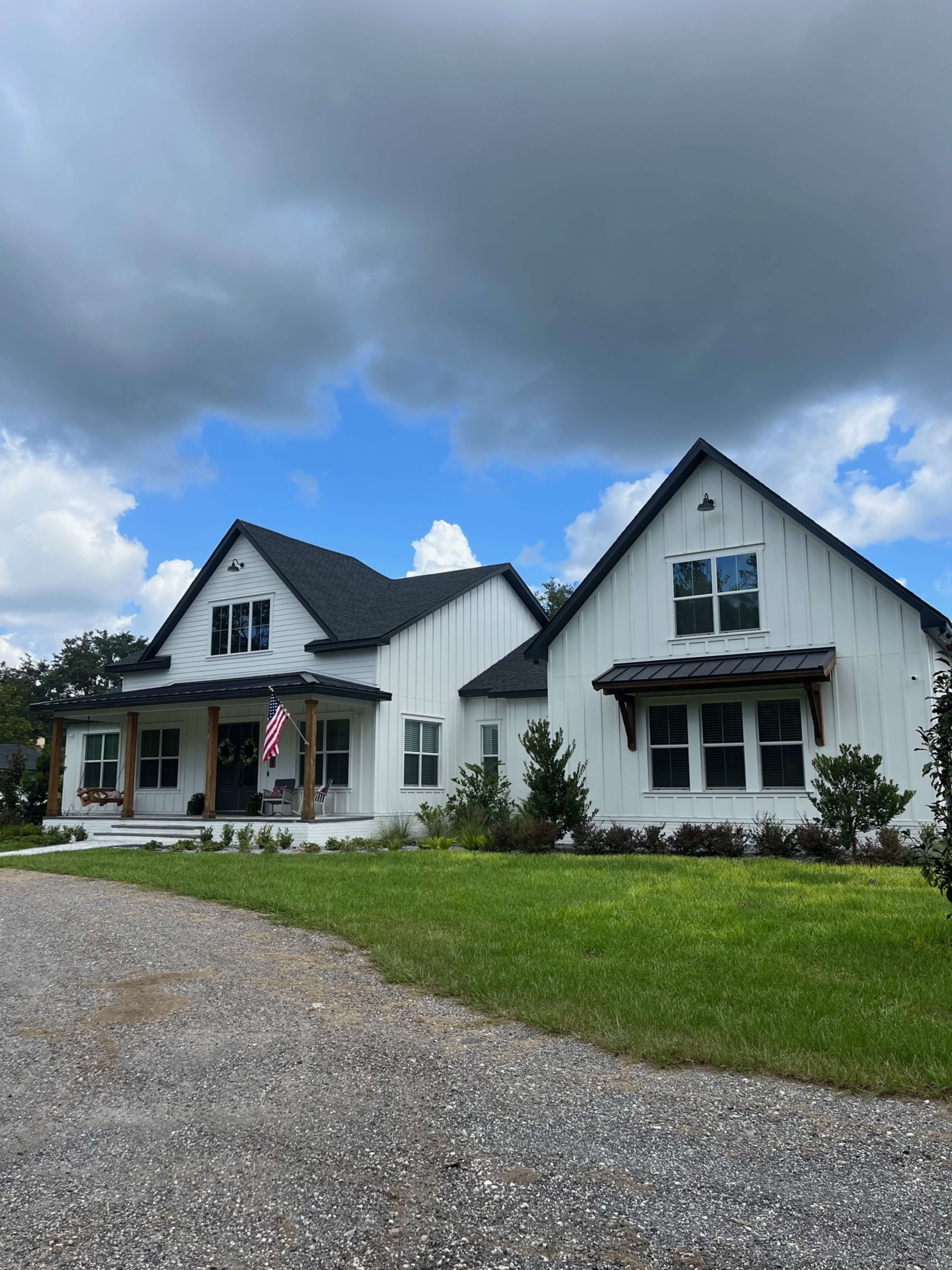 A two-story white house with a dark roof and an American flag in front is surrounded by grass and shrubs under a partly cloudy sky.