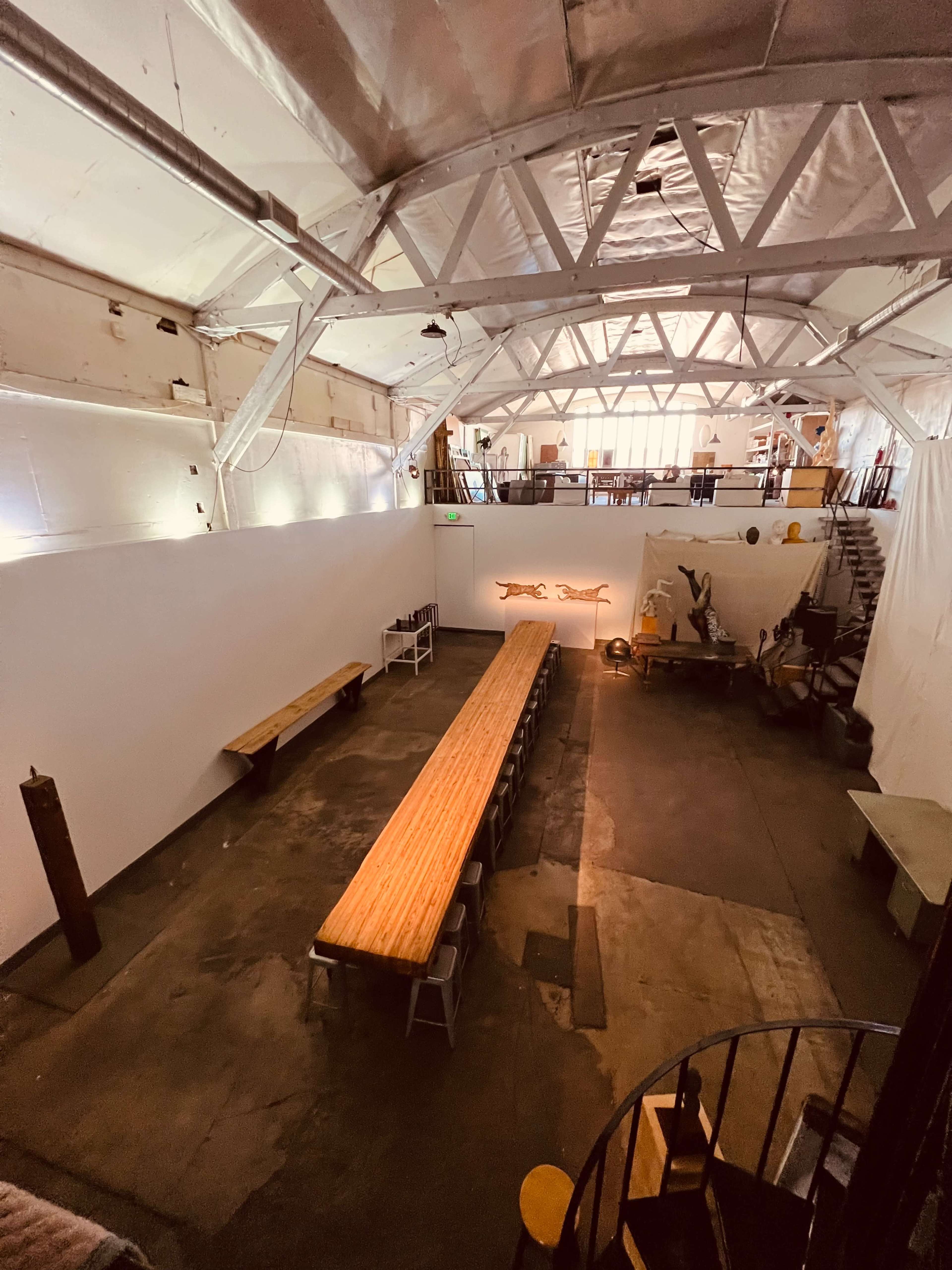 The image shows a spacious, industrial-style room with a long wooden dining table and a staircase on one side.