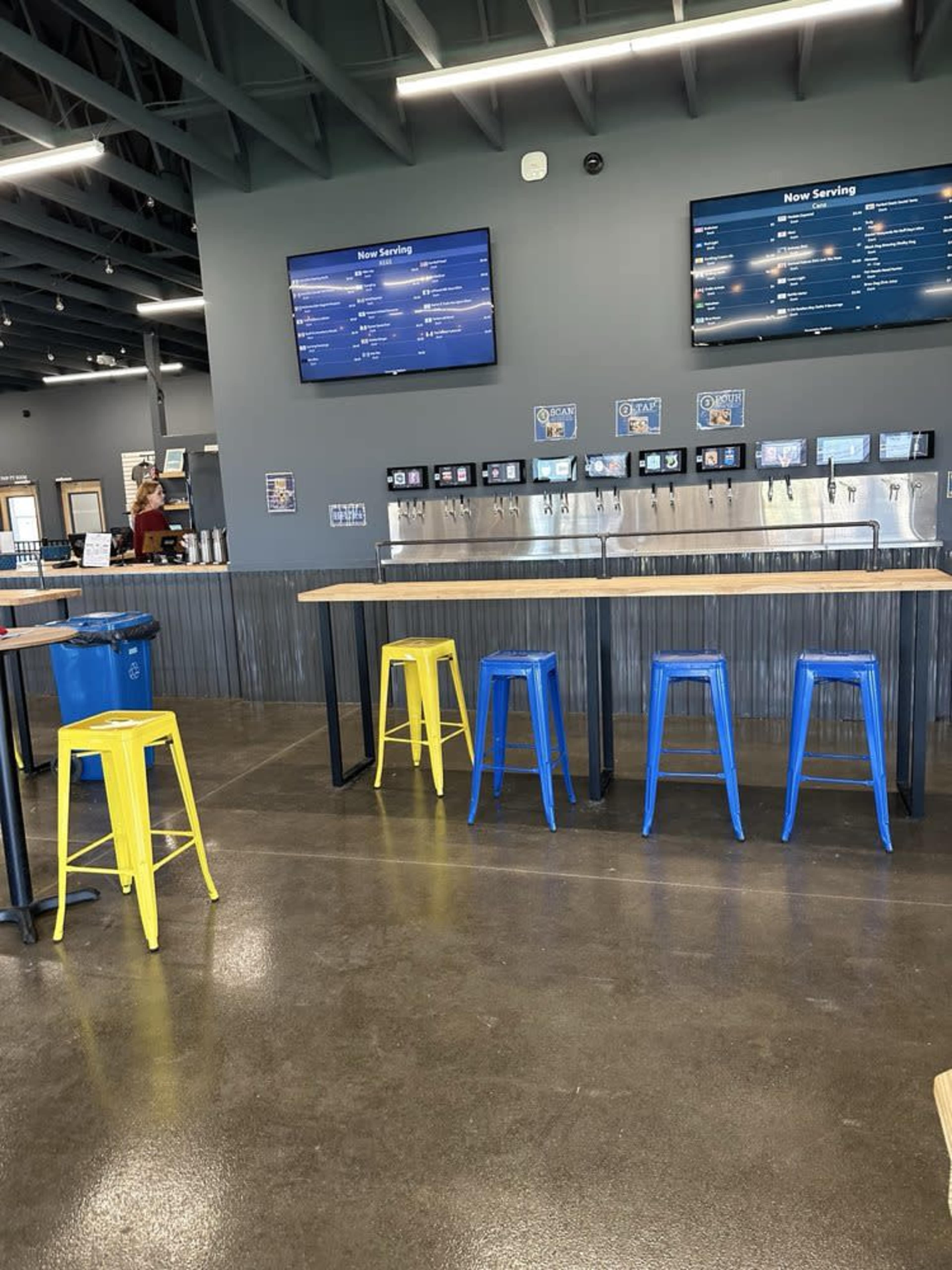 The image shows a modern beverage taproom with a bar counter, colorful stools, and digital menu screens on the wall.