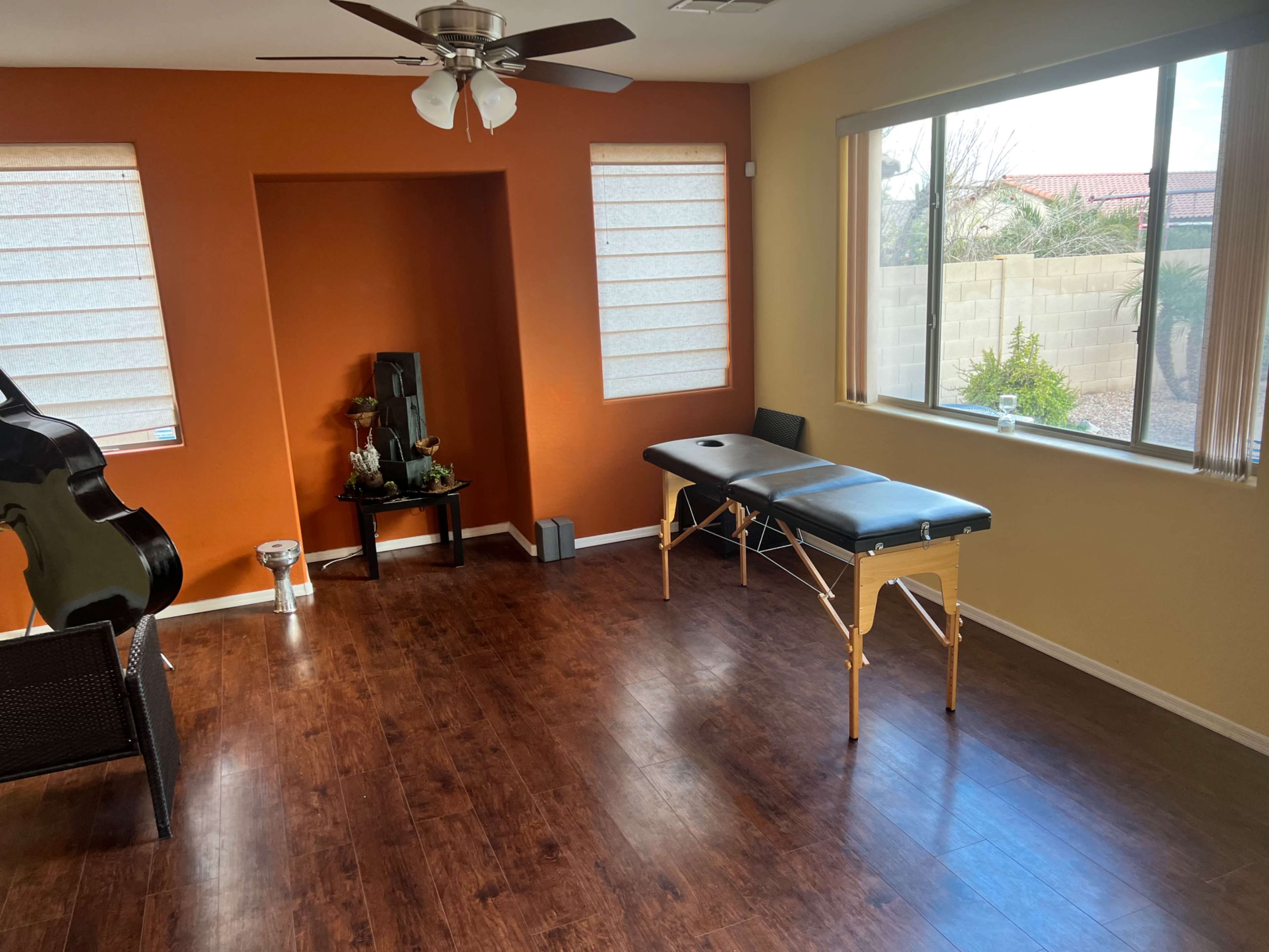 The room features brown wood flooring, an orange accent wall, a massage table, a black chair, and large windows providing natural light.