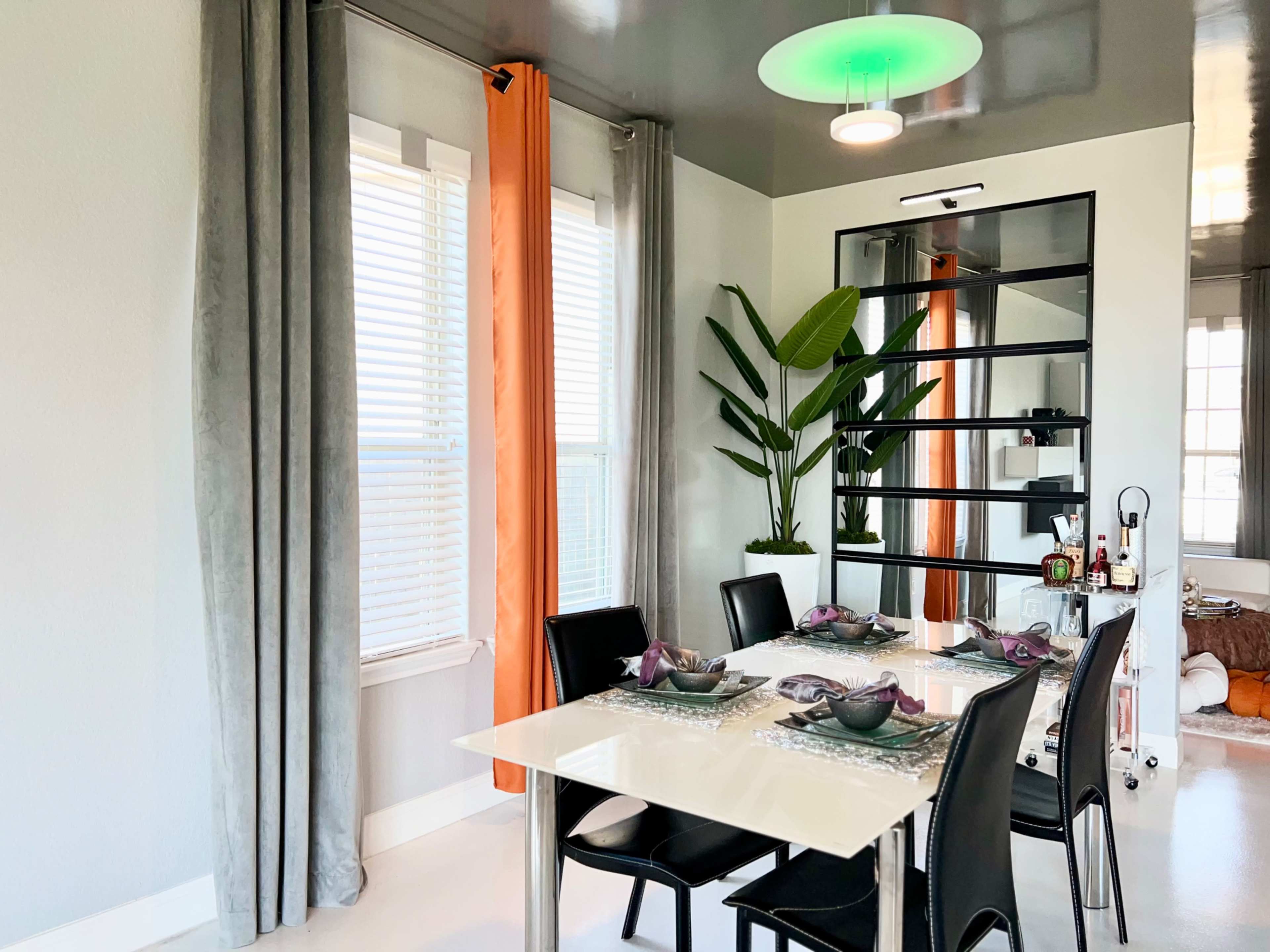 A modern dining area features a glass table surrounded by black chairs, with large windows adorned with gray and orange curtains and a tall plant in the corner.