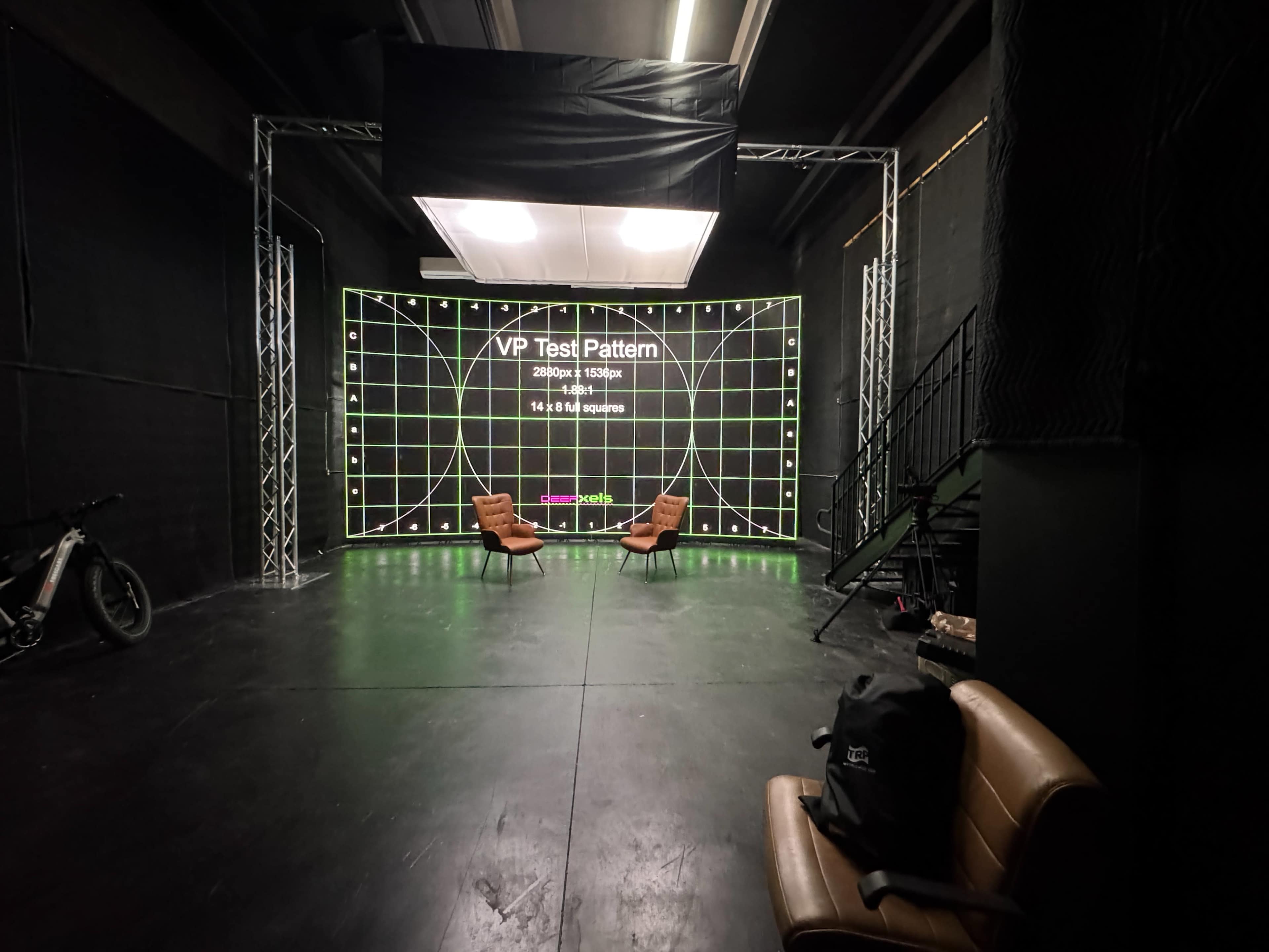 The image shows a modern studio space with a large display wall featuring a grid test pattern, two brown chairs facing the screen, and a bicycle in the corner.