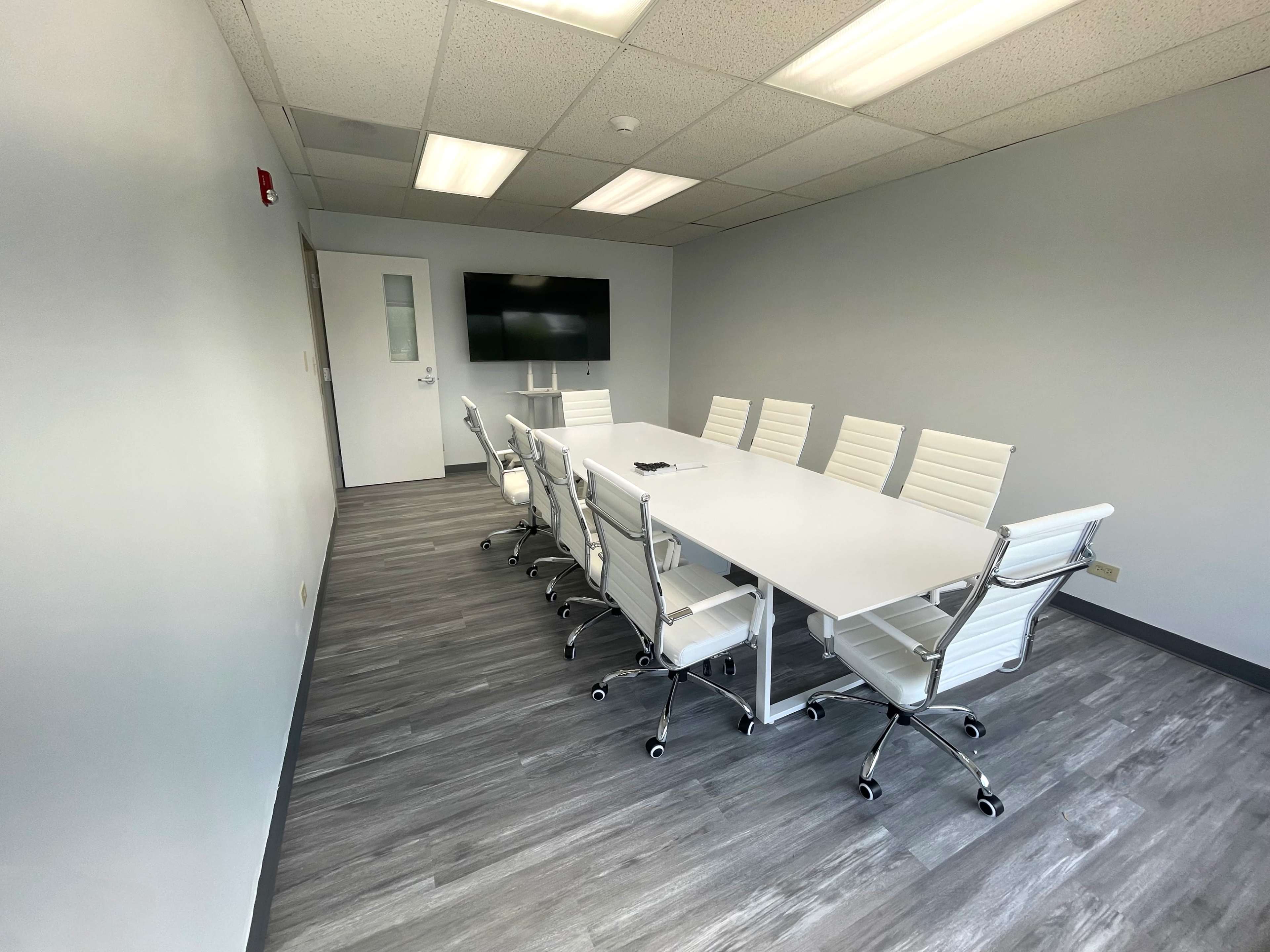 A sleek conference room features a long white table surrounded by eight white rolling chairs and a wall-mounted screen.