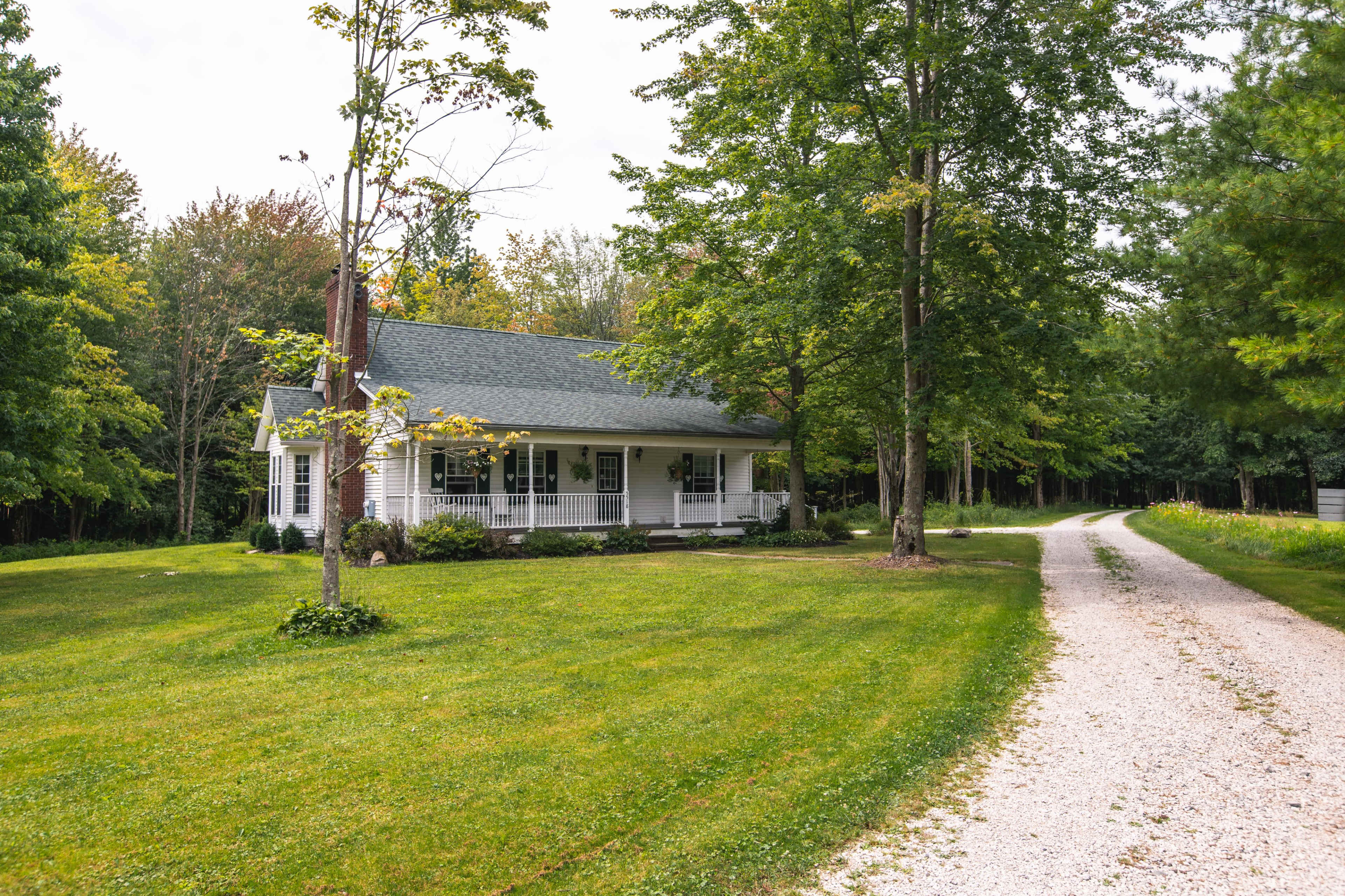 A gravel driveway leads to a one-story house surrounded by green grass and trees.