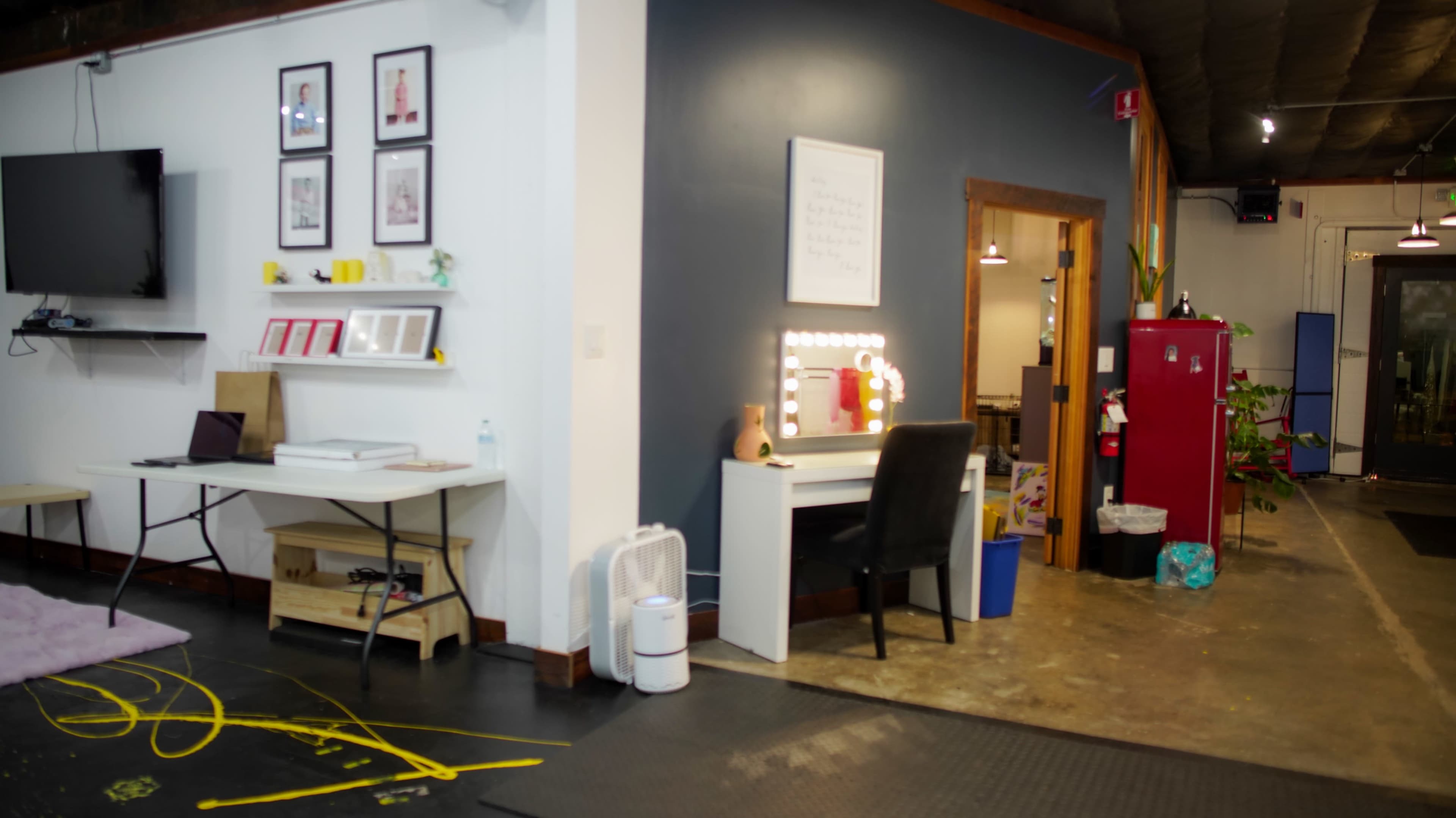 The image shows a room divided into two areas: one side features a desk with framed pictures on the wall, and the other side contains a vanity with a chair and a red storage unit.