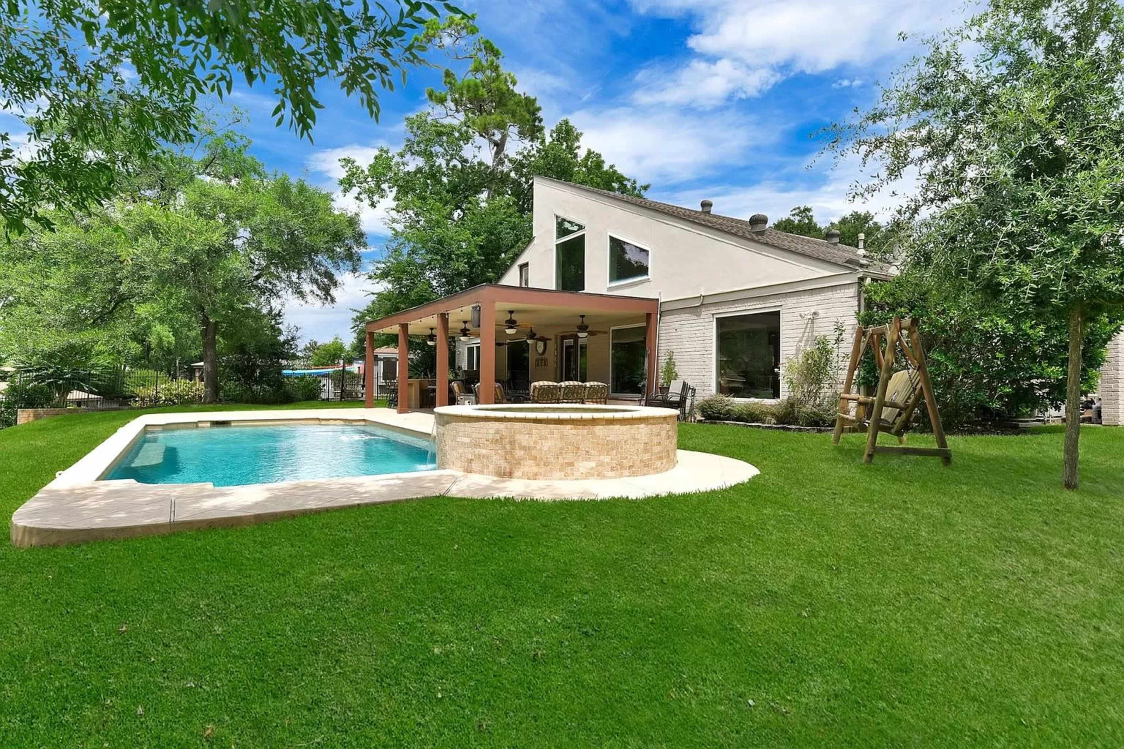 A backyard with a swimming pool, a covered patio area, and a swing set, framed by trees and a well-maintained lawn.