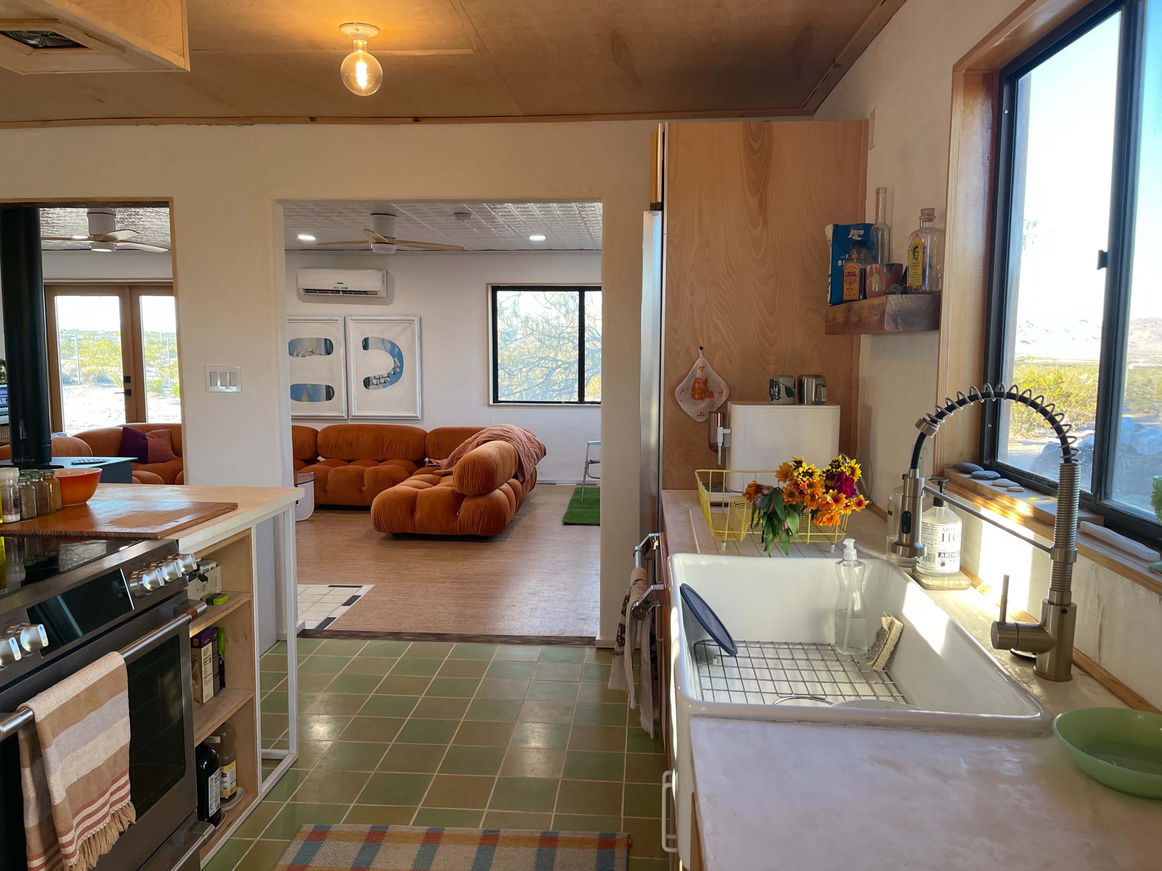 A modern kitchen features a sink and stove with wooden cabinetry, leading to a bright open living area with orange couches and large windows.