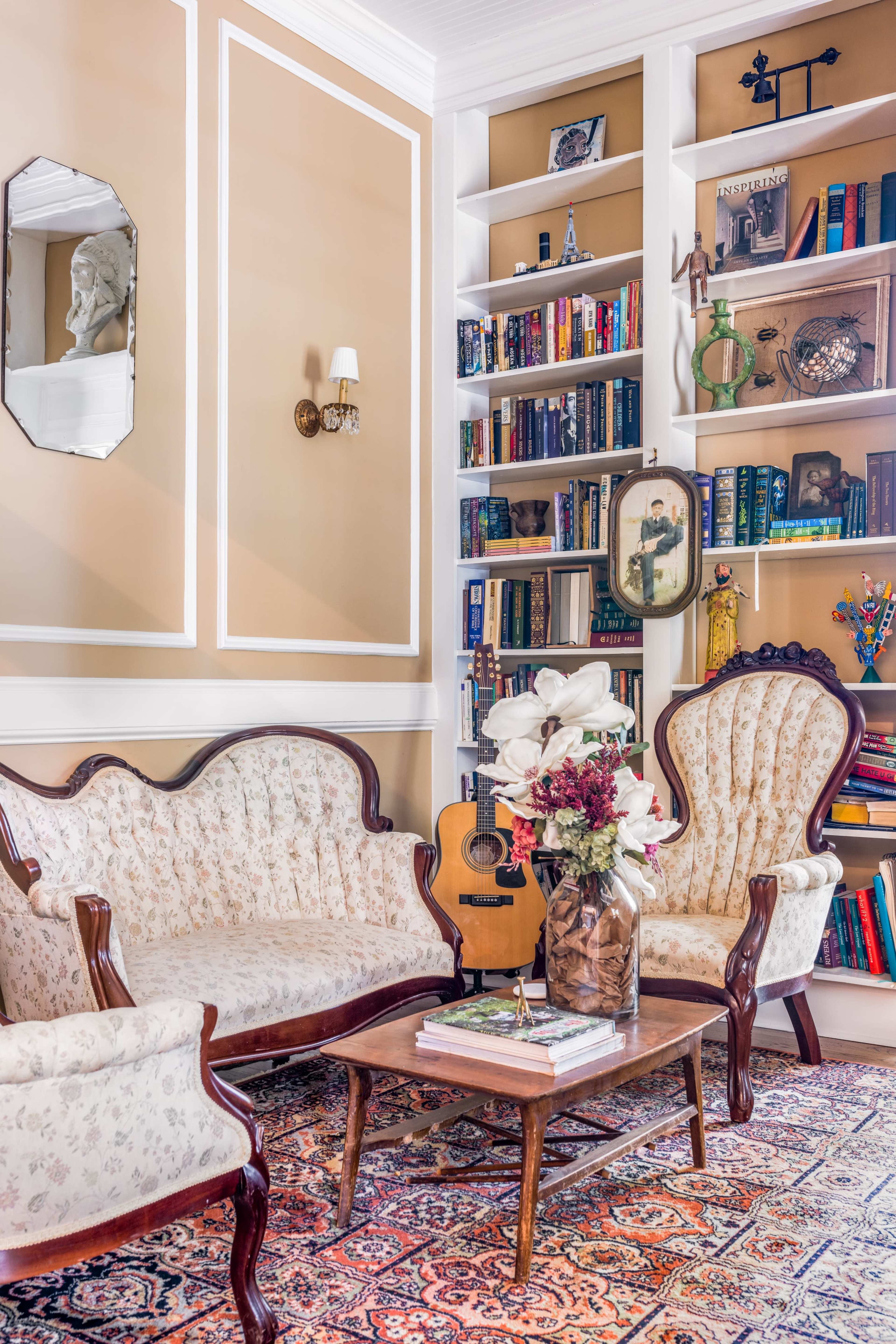A cozy living room features two upholstered armchairs and a matching sofa surrounding a low wooden coffee table, complemented by a bookshelf filled with books and decorative items.