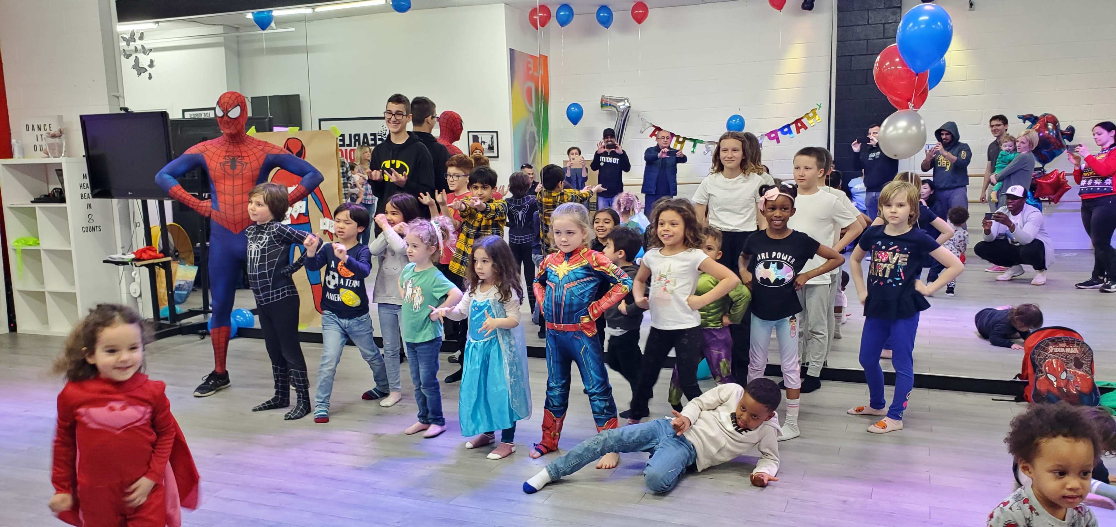 A group of children dressed in various superhero costumes gathers in a dance studio decorated with balloons and streamers.
