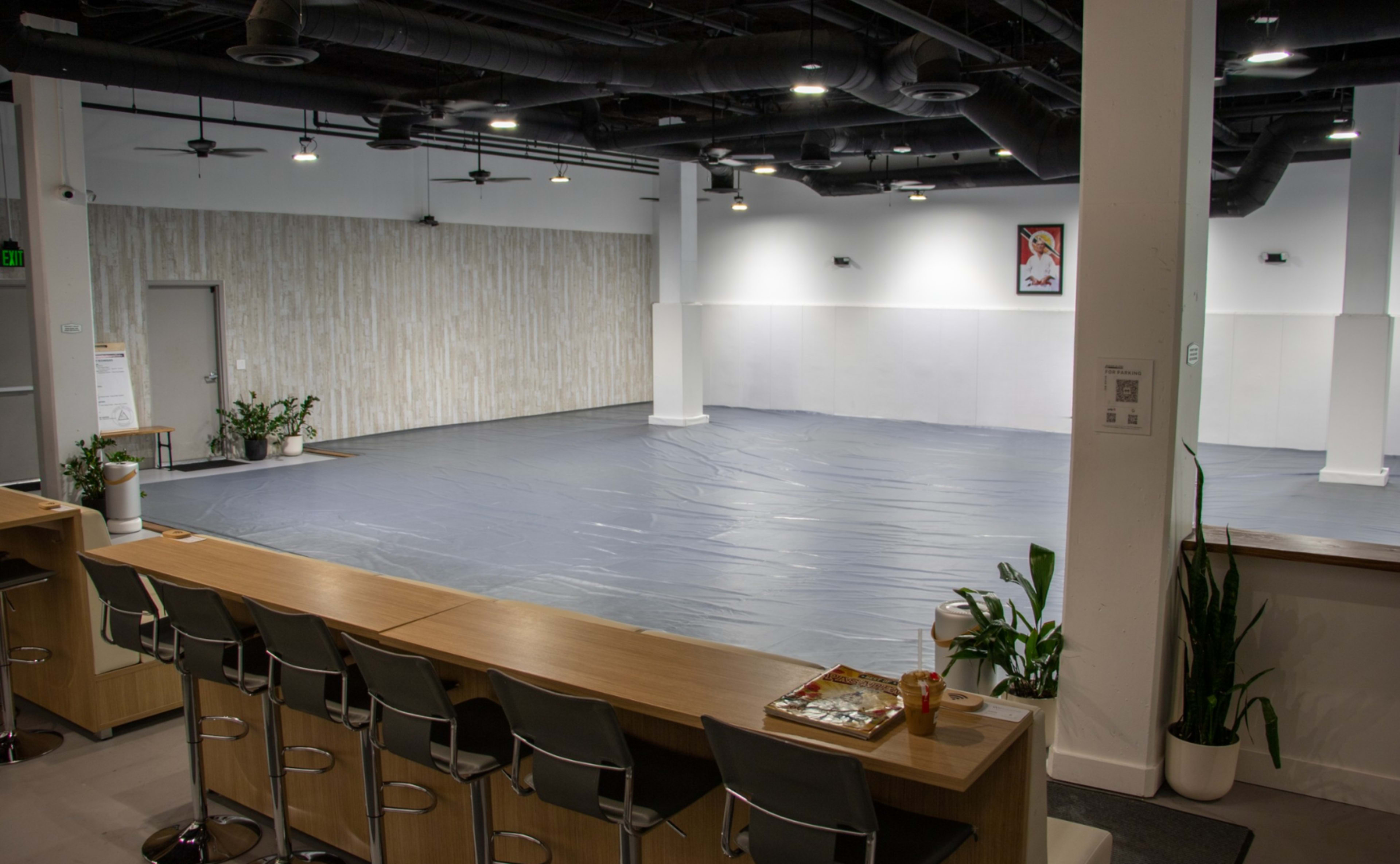 The image shows a spacious indoor area with a gray floor covering, surrounded by white walls and minimal decor, featuring a seating area lined with bar stools and plants.