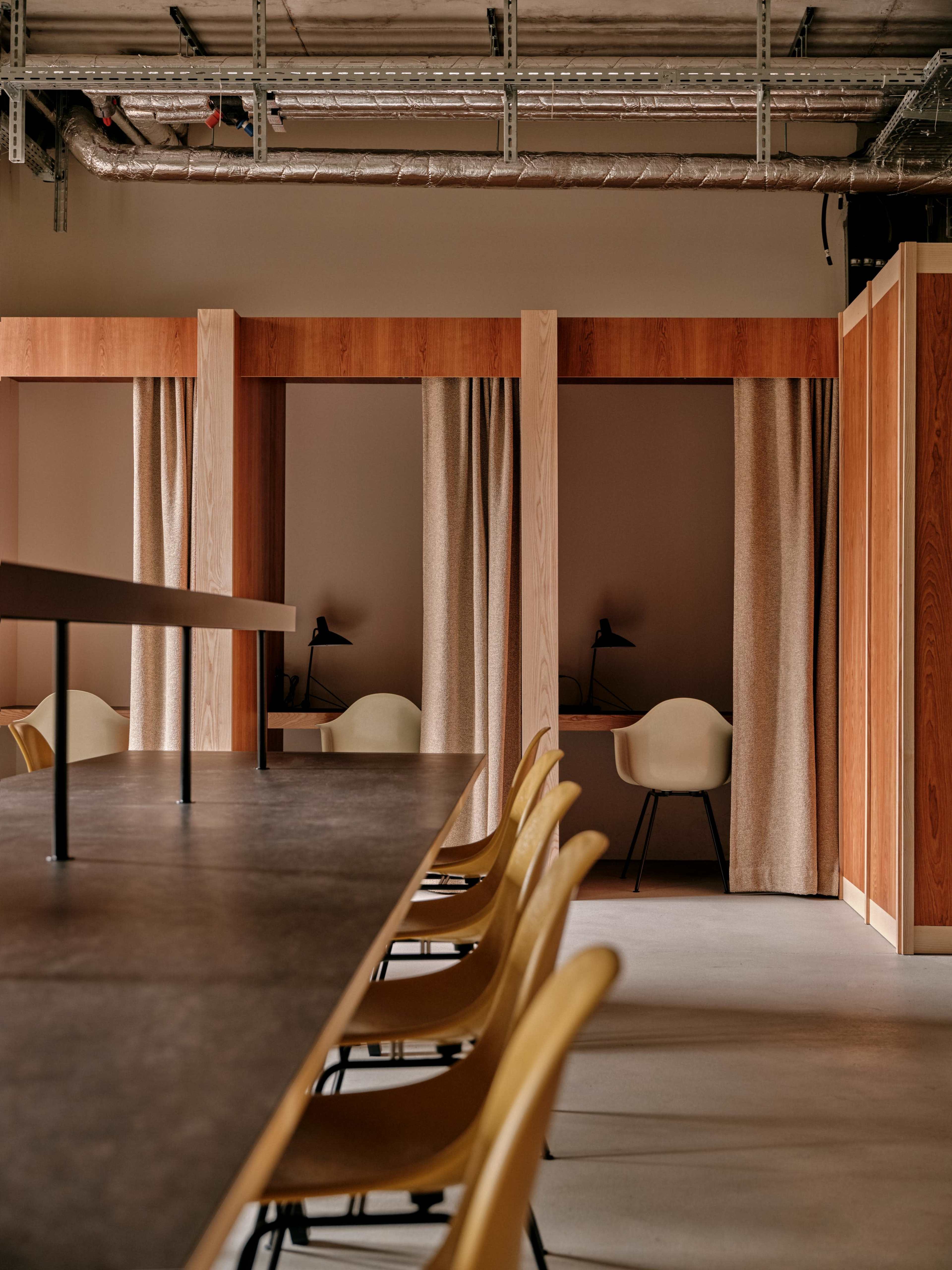 A modern workspace featuring a long table with yellow chairs and private cubicles separated by curtains in a minimalist setting.
