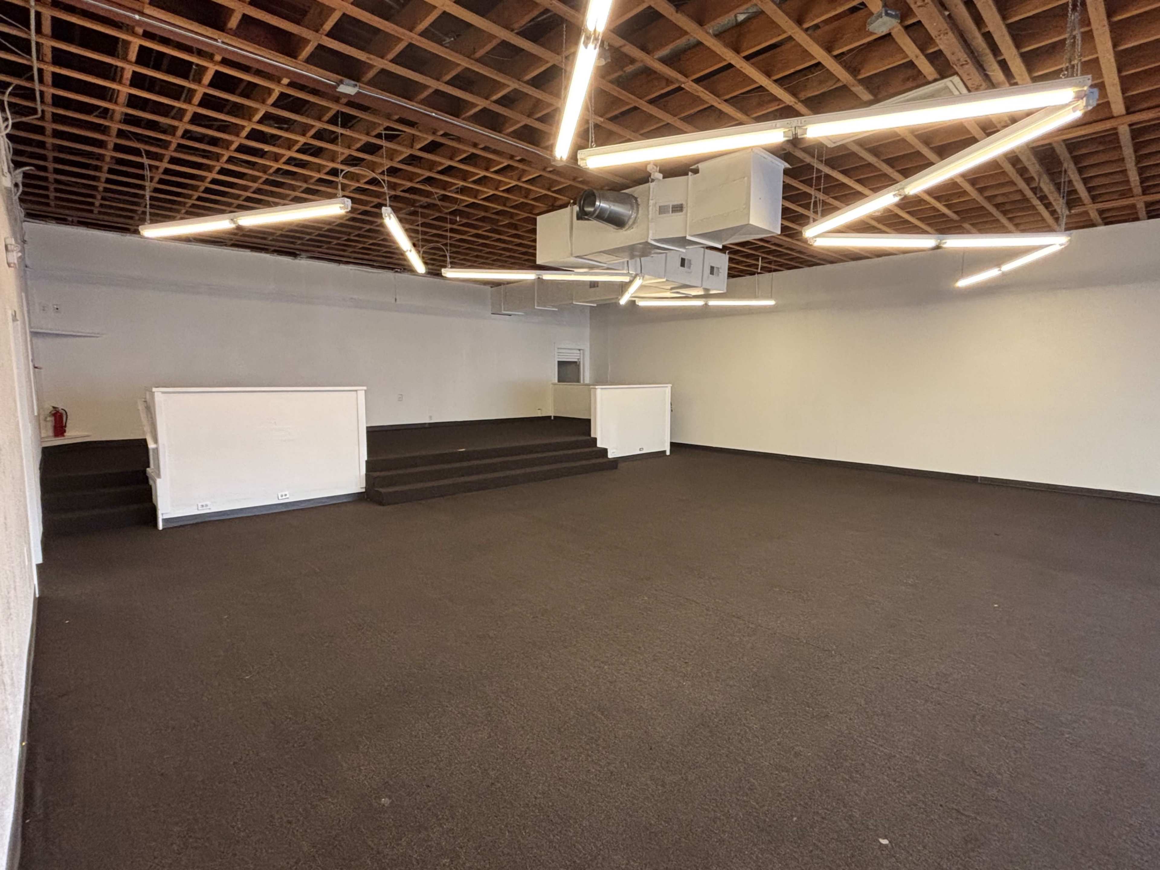 The image shows an empty room with a dark carpet, white walls, a raised stage area, and modern overhead lighting.