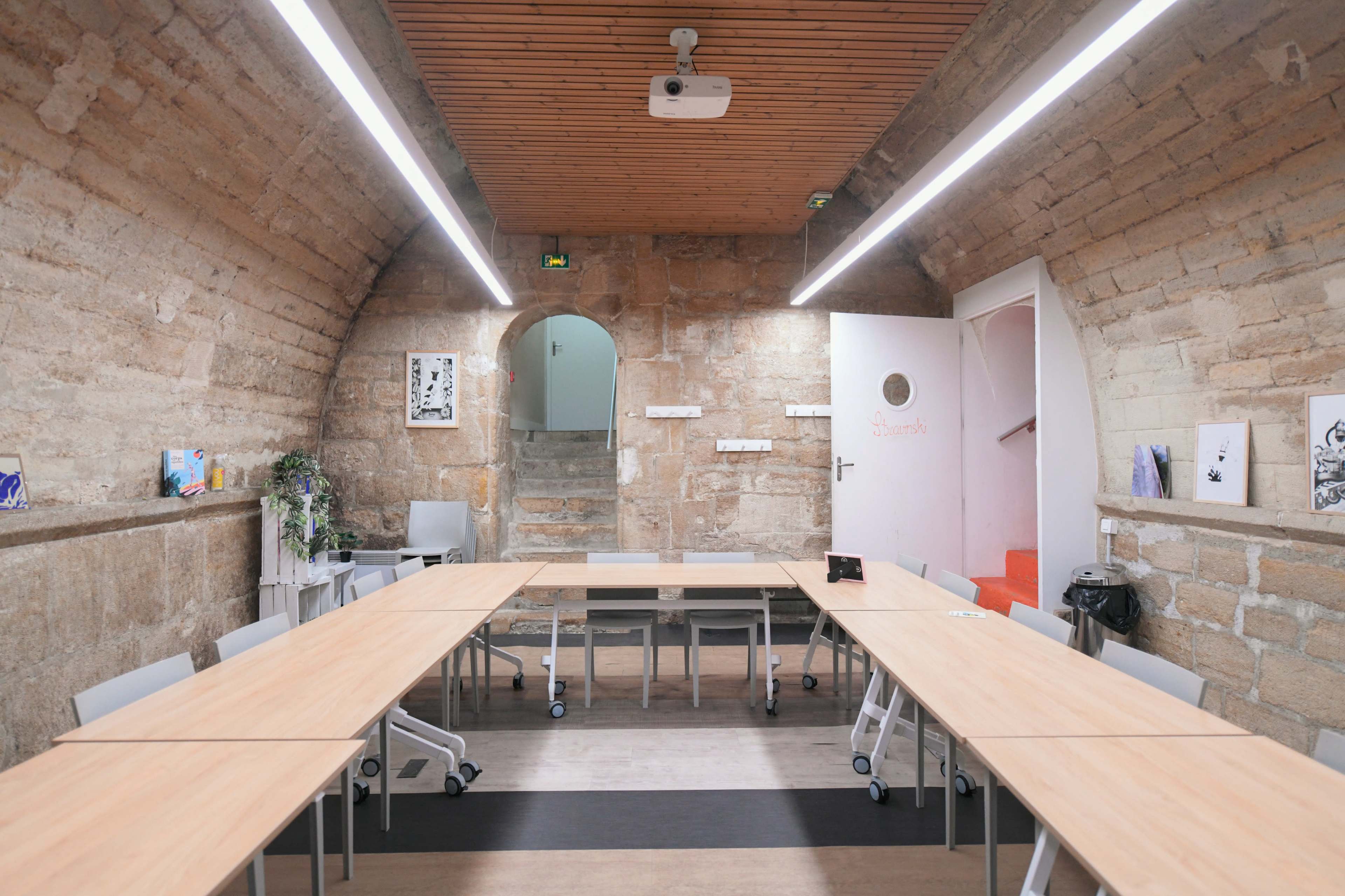 An arched room with stone walls, featuring two long wooden tables arranged in a U-shape and a staircase leading to another area.