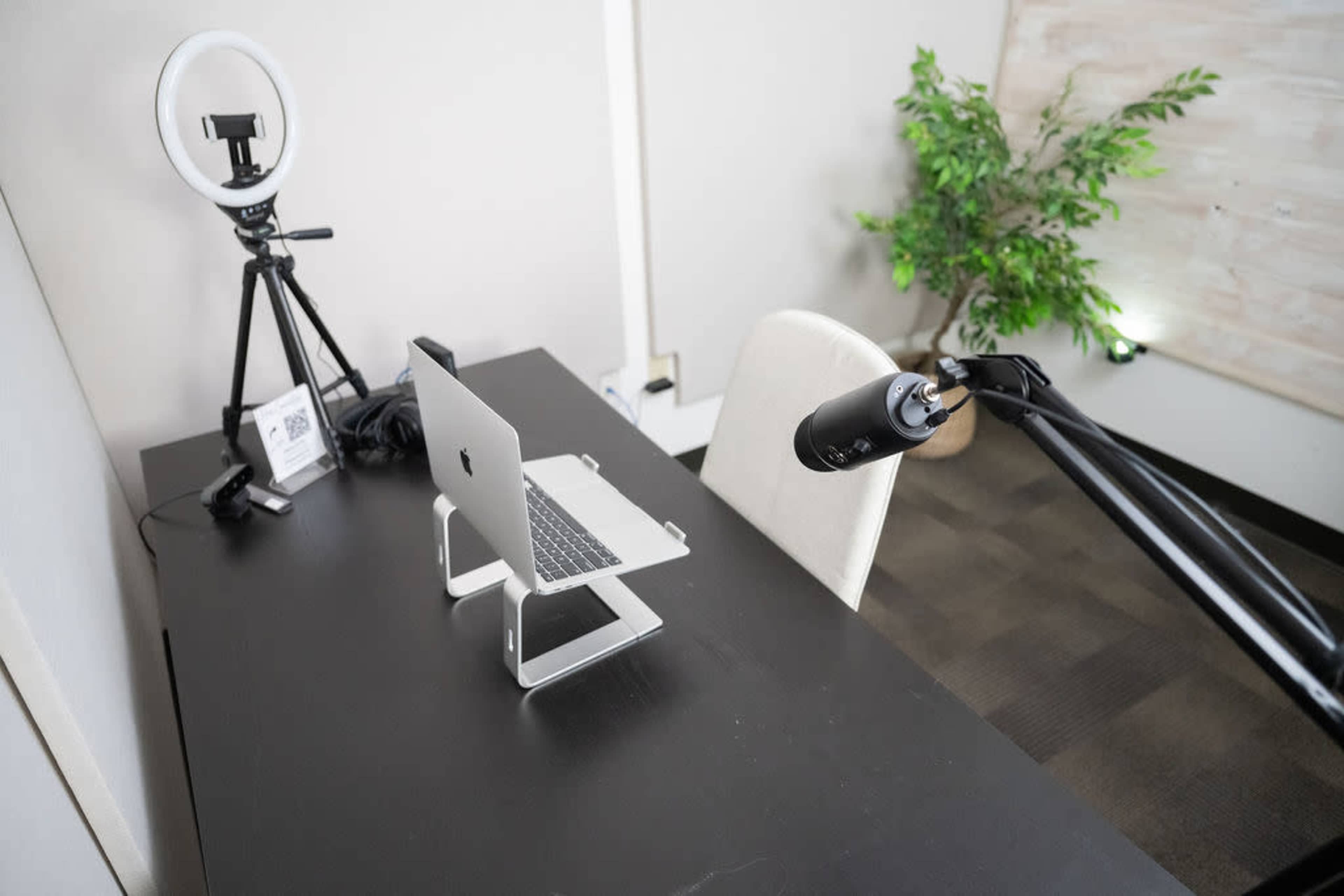 A workspace featuring a laptop on a stand, a microphone on an adjustable arm, a ring light on a tripod, and a potted plant in the background.