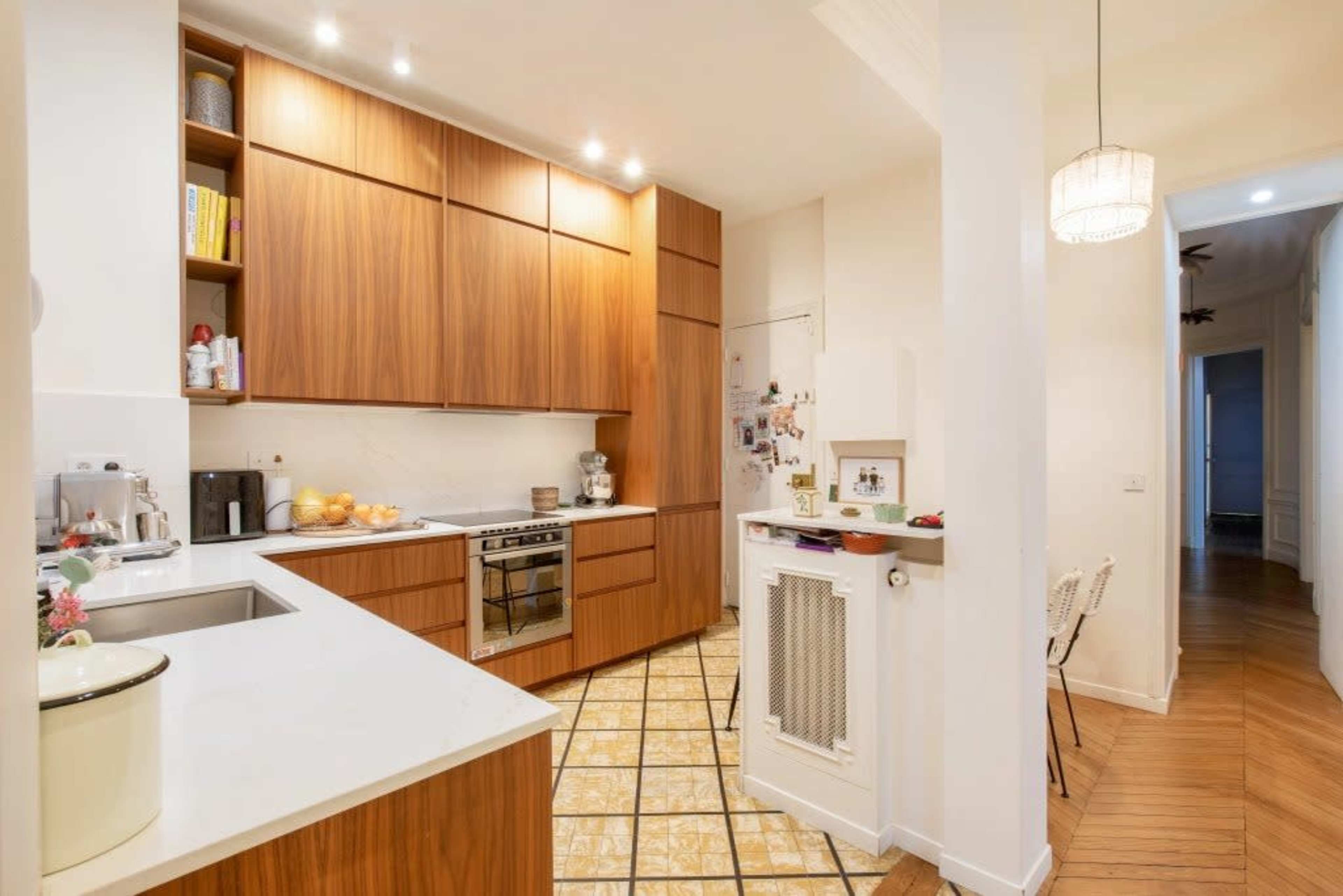 The image shows a modern kitchen with wooden cabinetry, a white countertop, and a tiled floor, adjacent to a hallway.