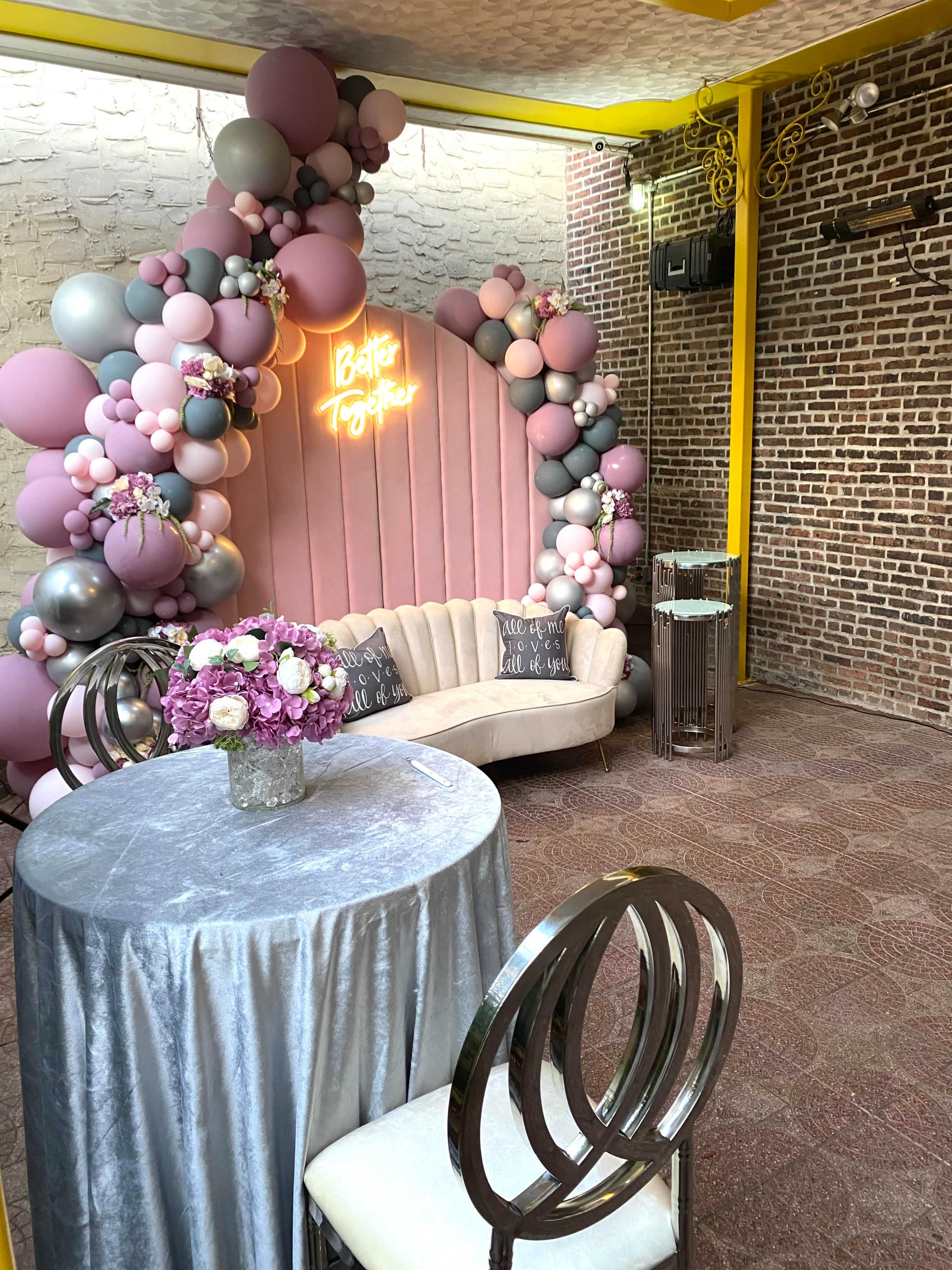 The image shows a decorated corner of a venue featuring a beige couch, a round table covered with a silver tablecloth, and a colorful balloon arch with a neon sign that reads "Better Together."