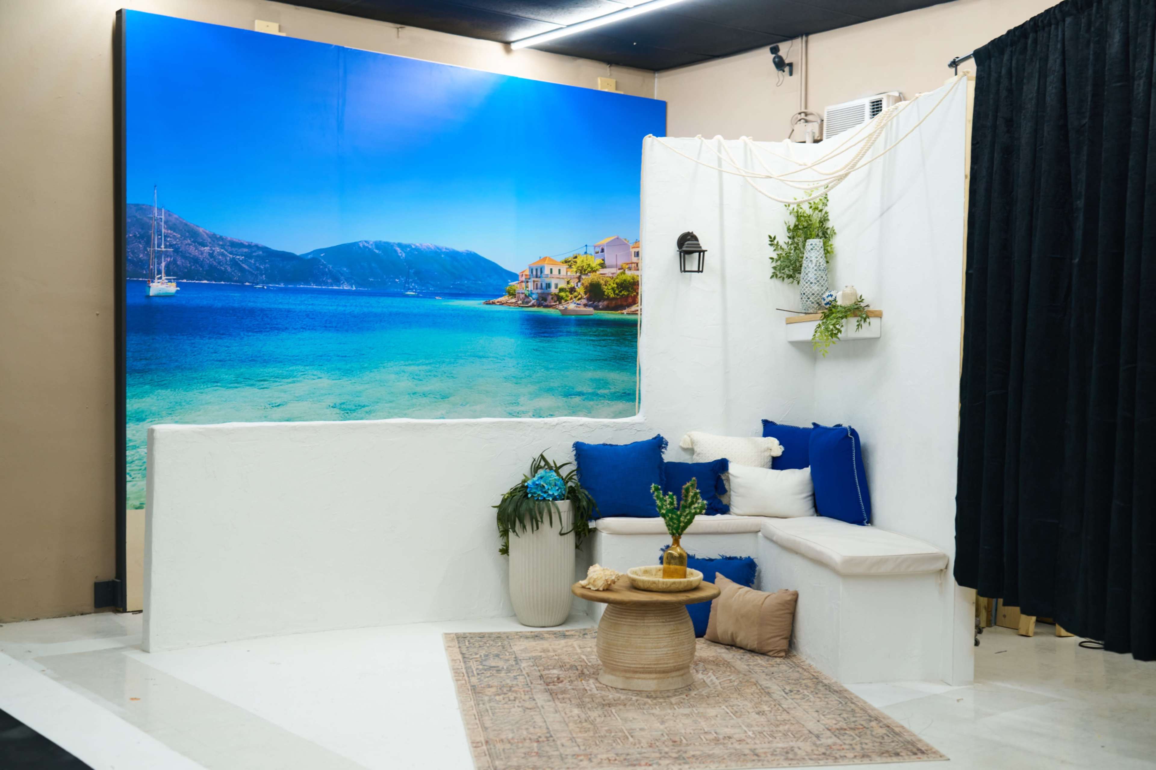 The image shows a stylized indoor setting featuring a white backdrop with a coastal landscape mural, a seating area with blue cushions, a decorative table, and potted plants.