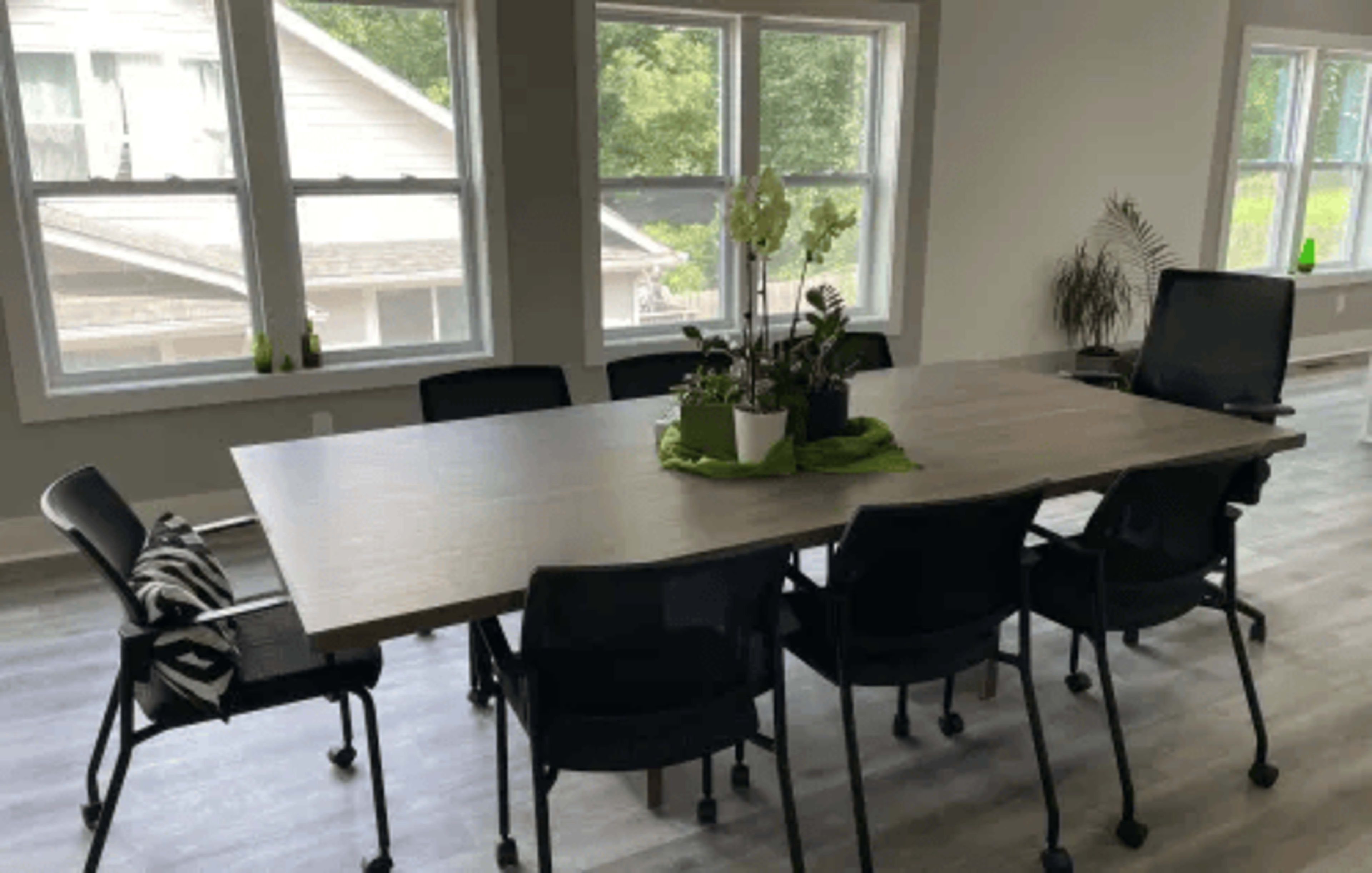 A large rectangular table surrounded by black chairs, with potted plants in the center, set in a brightly lit room with multiple large windows.