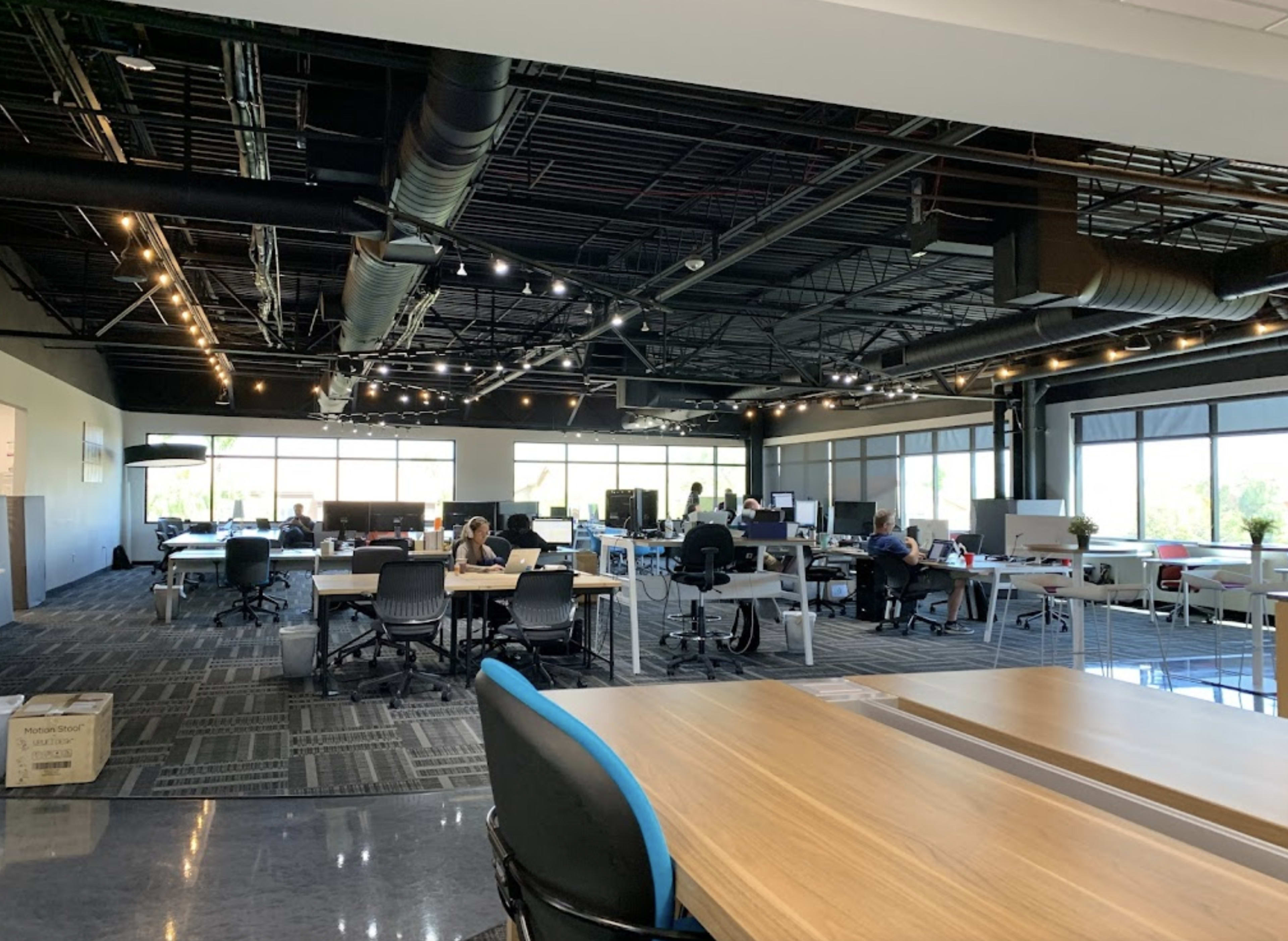 A spacious office environment features multiple workstations, large windows, and several employees engaged in various tasks.