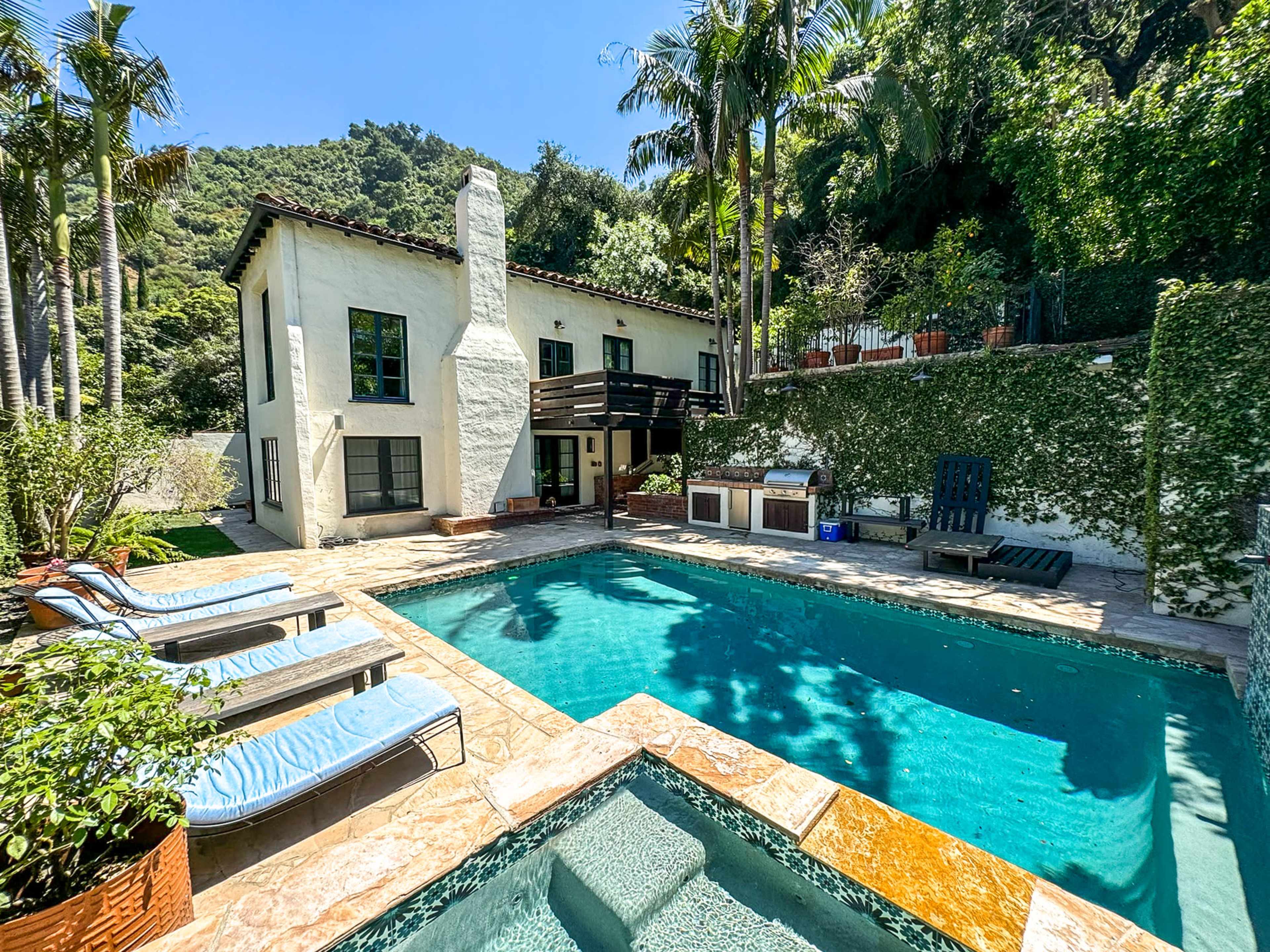 A two-story house with a swimming pool and outdoor seating area is surrounded by lush greenery and palm trees.
