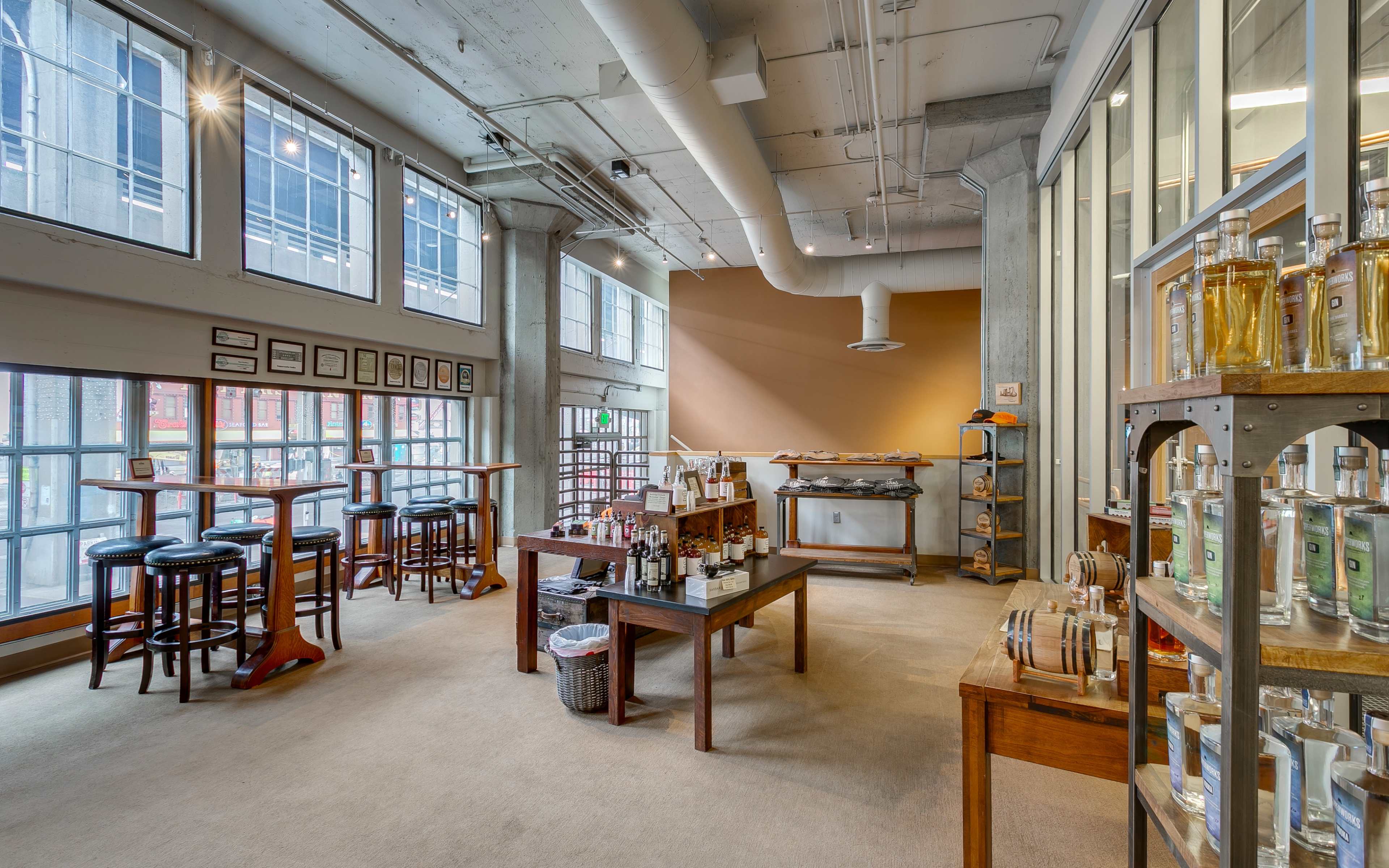 A spacious interior of a tasting room with wooden tables, bar stools, and shelves displaying bottles, illuminated by natural light from large windows.