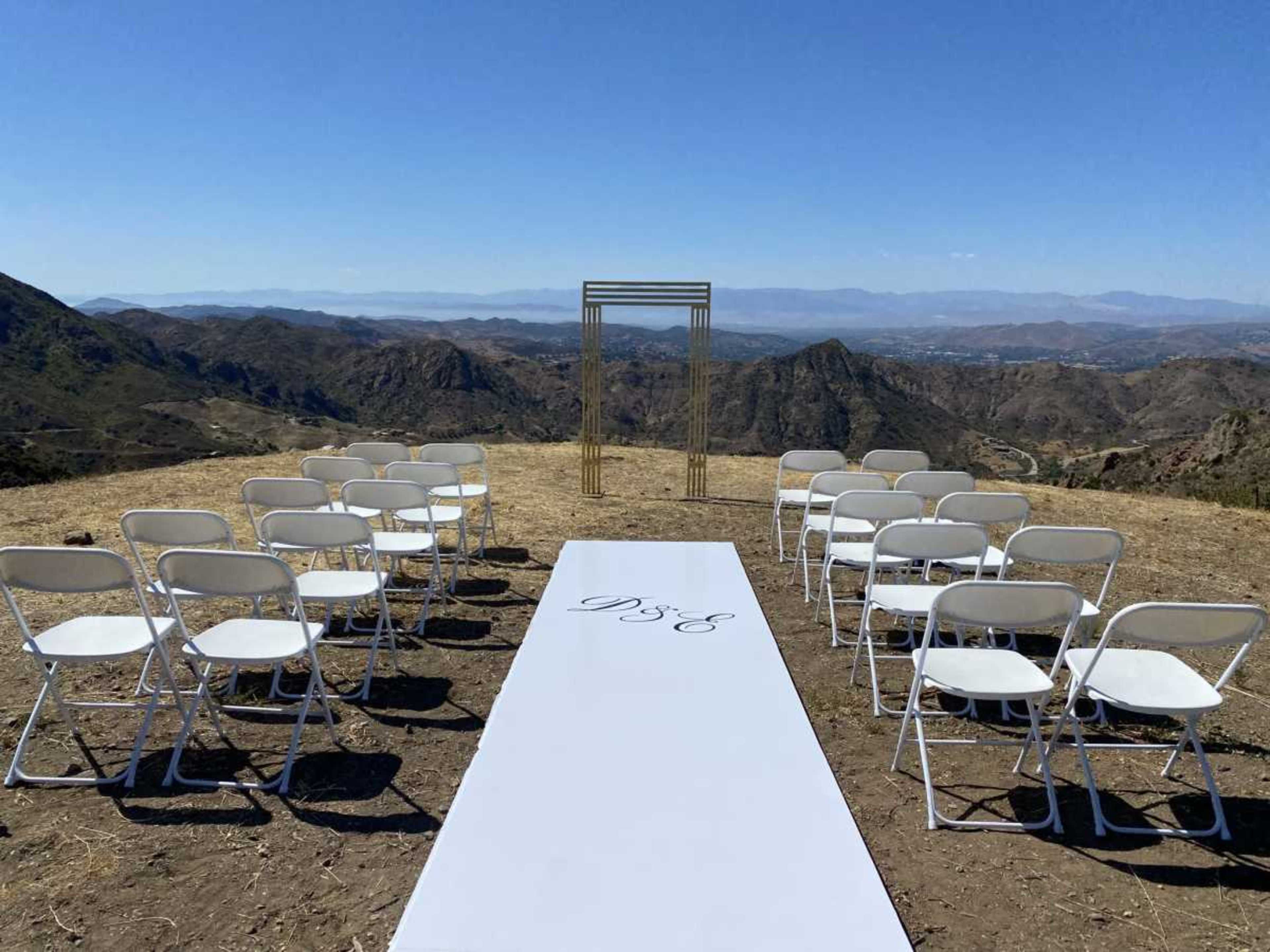 An outdoor wedding setup features rows of white chairs facing a decorated arch on a grassy area with a mountainous landscape in the background.