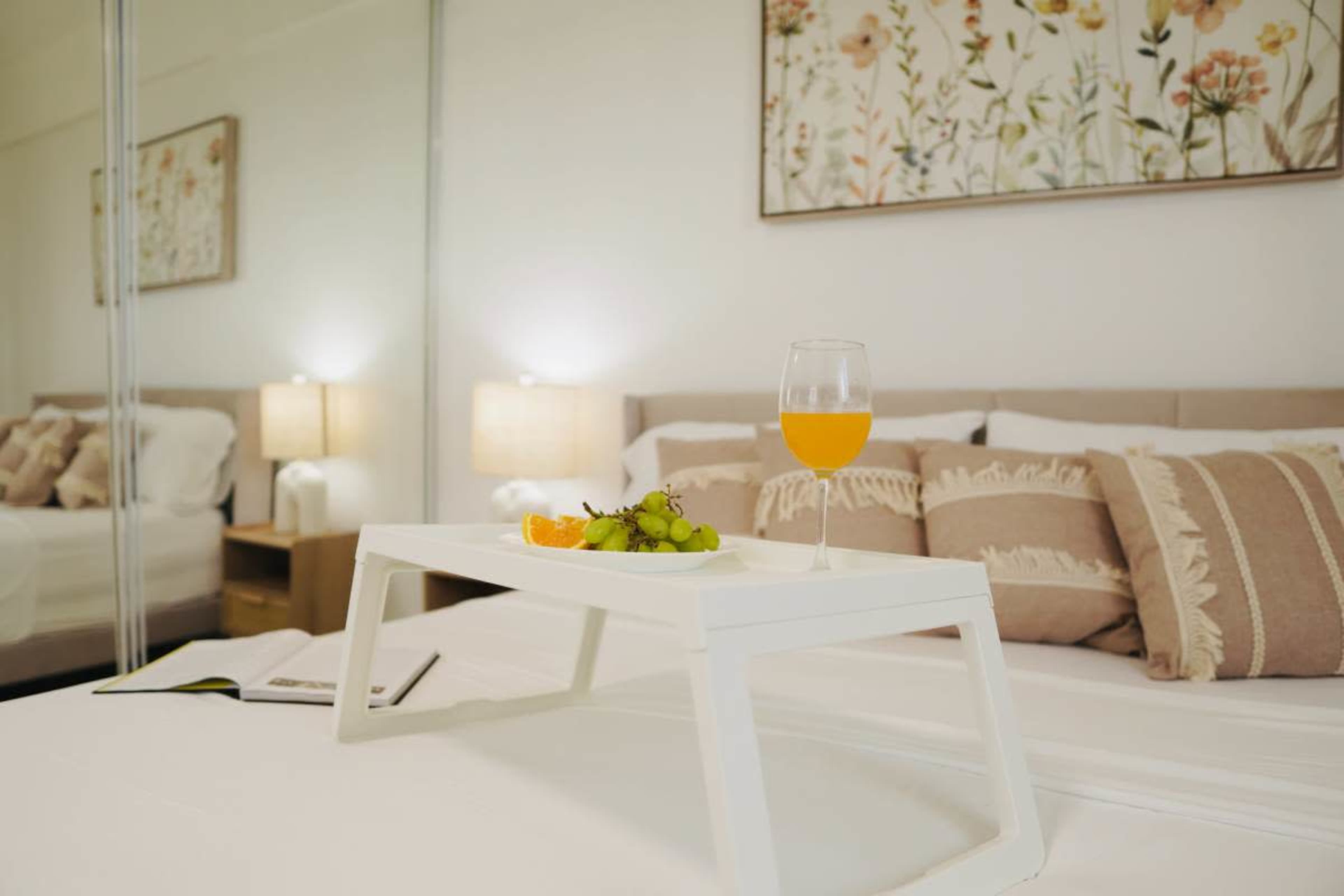 A tray with fruit and a drink is placed on a neatly made bed in a bright bedroom featuring floral artwork and mirrored closets.
