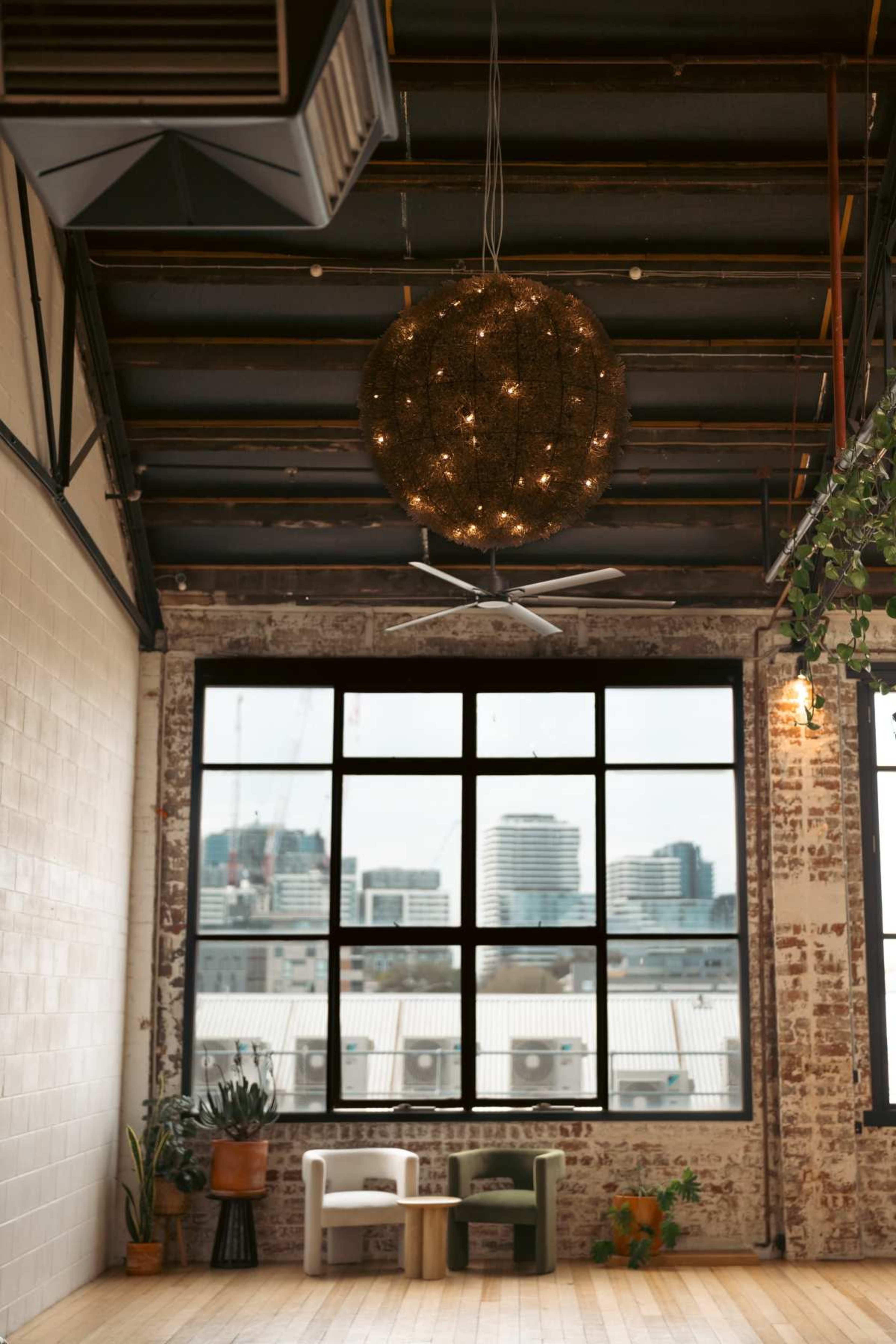 The image shows an industrial-style interior with a large, round pendant light, a ceiling fan, and a window revealing a city skyline in the background.