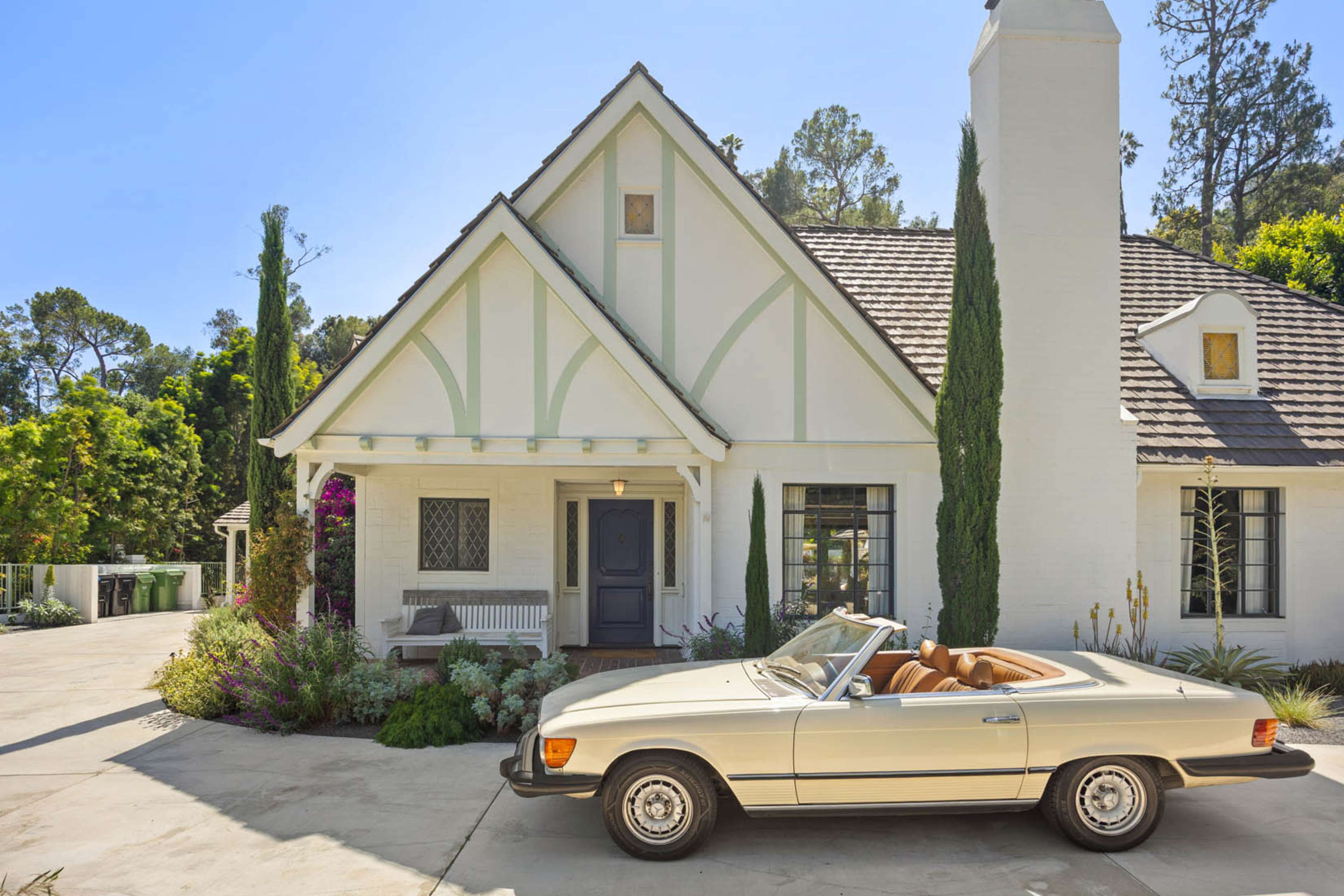 A vintage convertible car is parked in front of a white, gabled house surrounded by greenery.