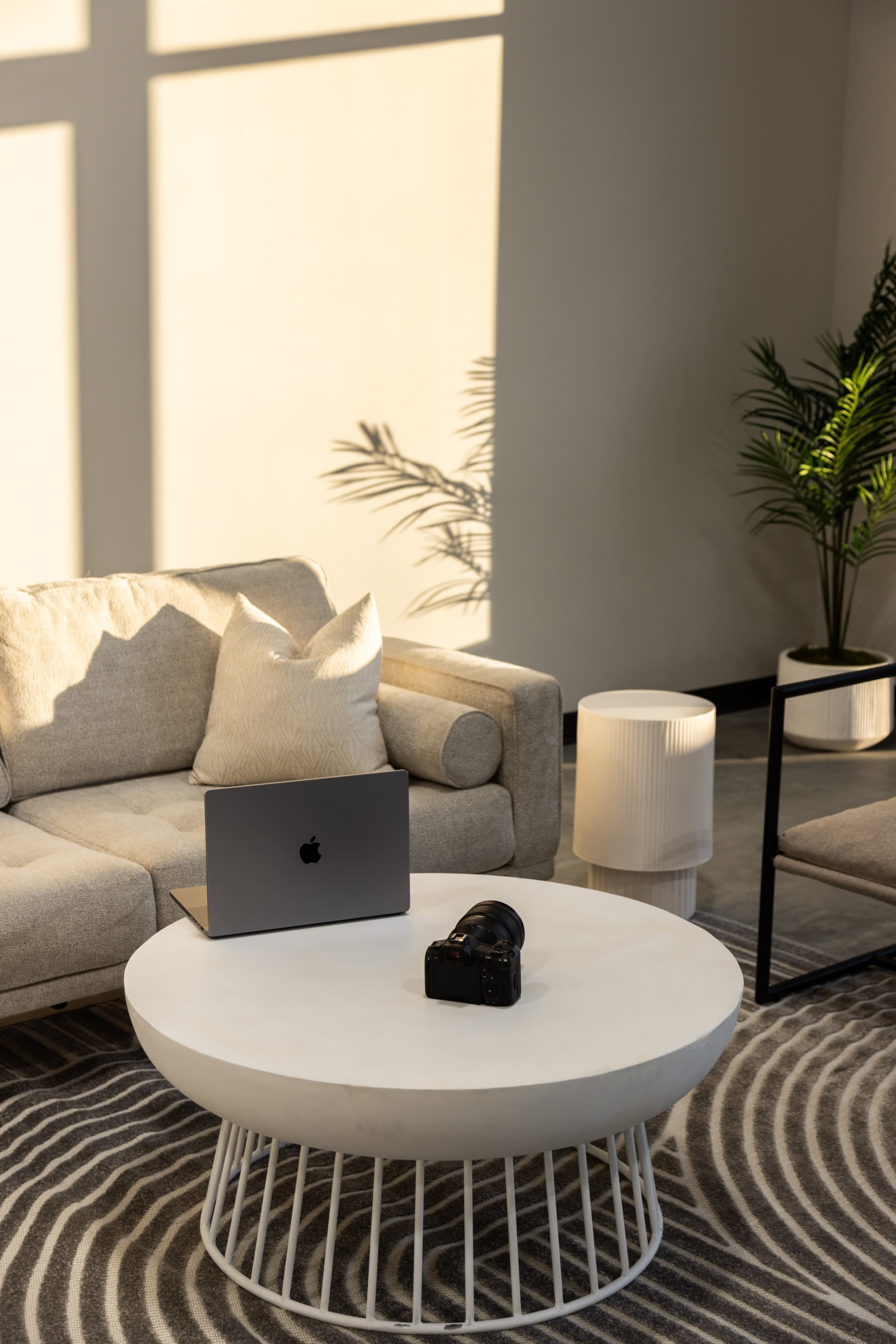 A laptop and camera are placed on a round coffee table in a softly lit living room with a light-colored sofa and decorative plants.