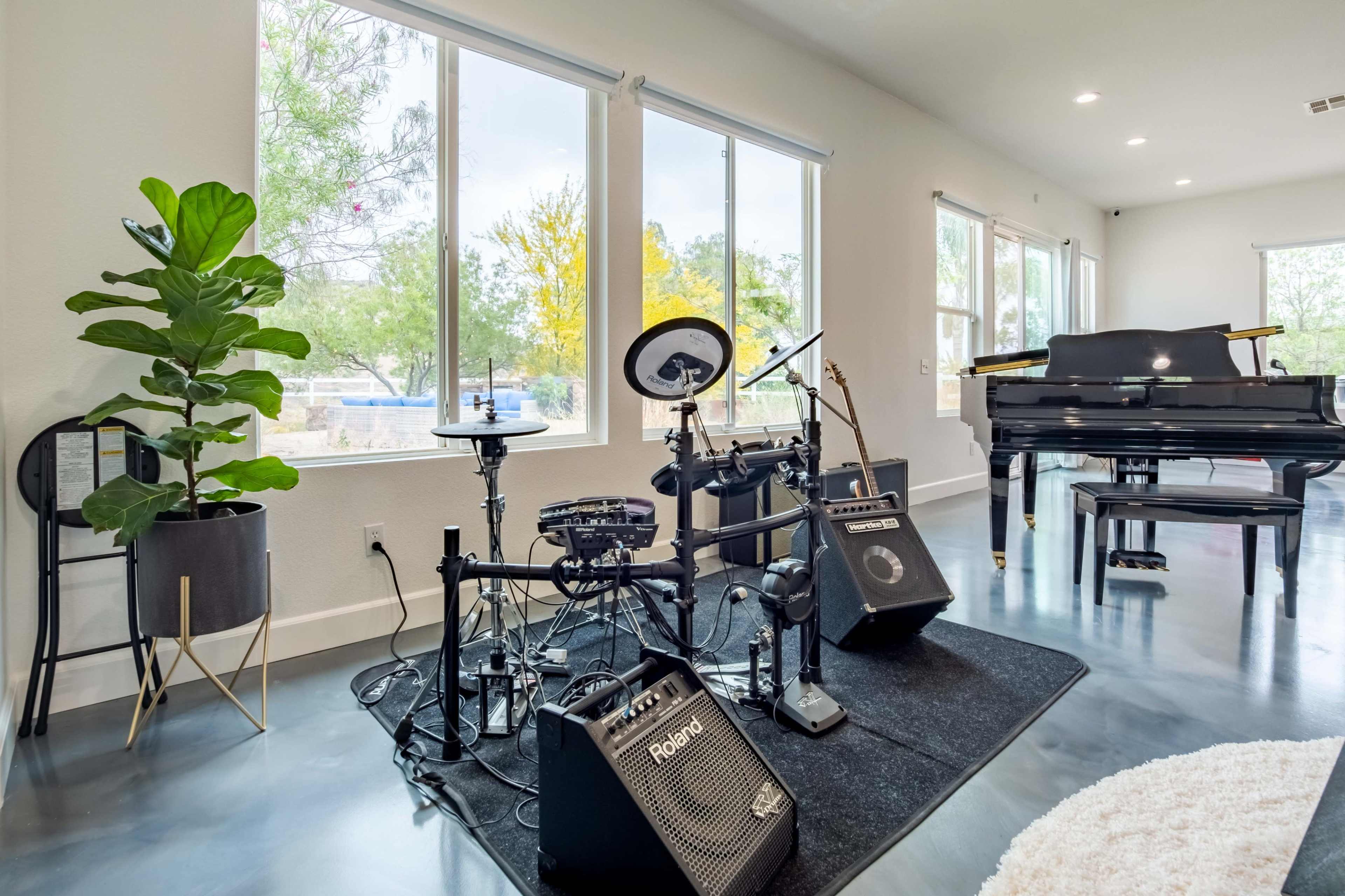 A music setup featuring an electronic drum kit, amplifiers, and a grand piano in a well-lit room with large windows.