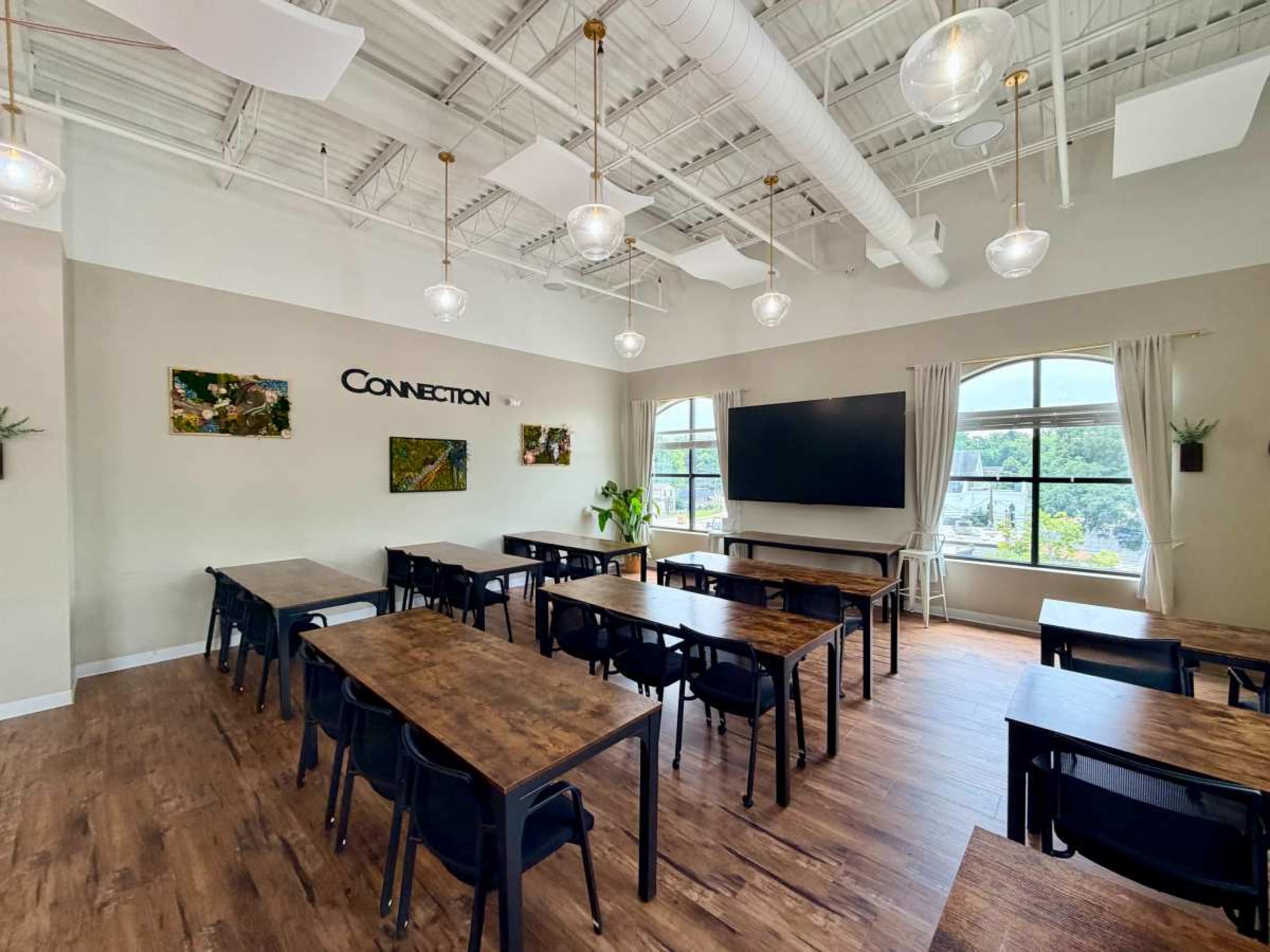 The image shows a modern classroom with several wooden tables, black chairs, large windows, and a wall-mounted screen.