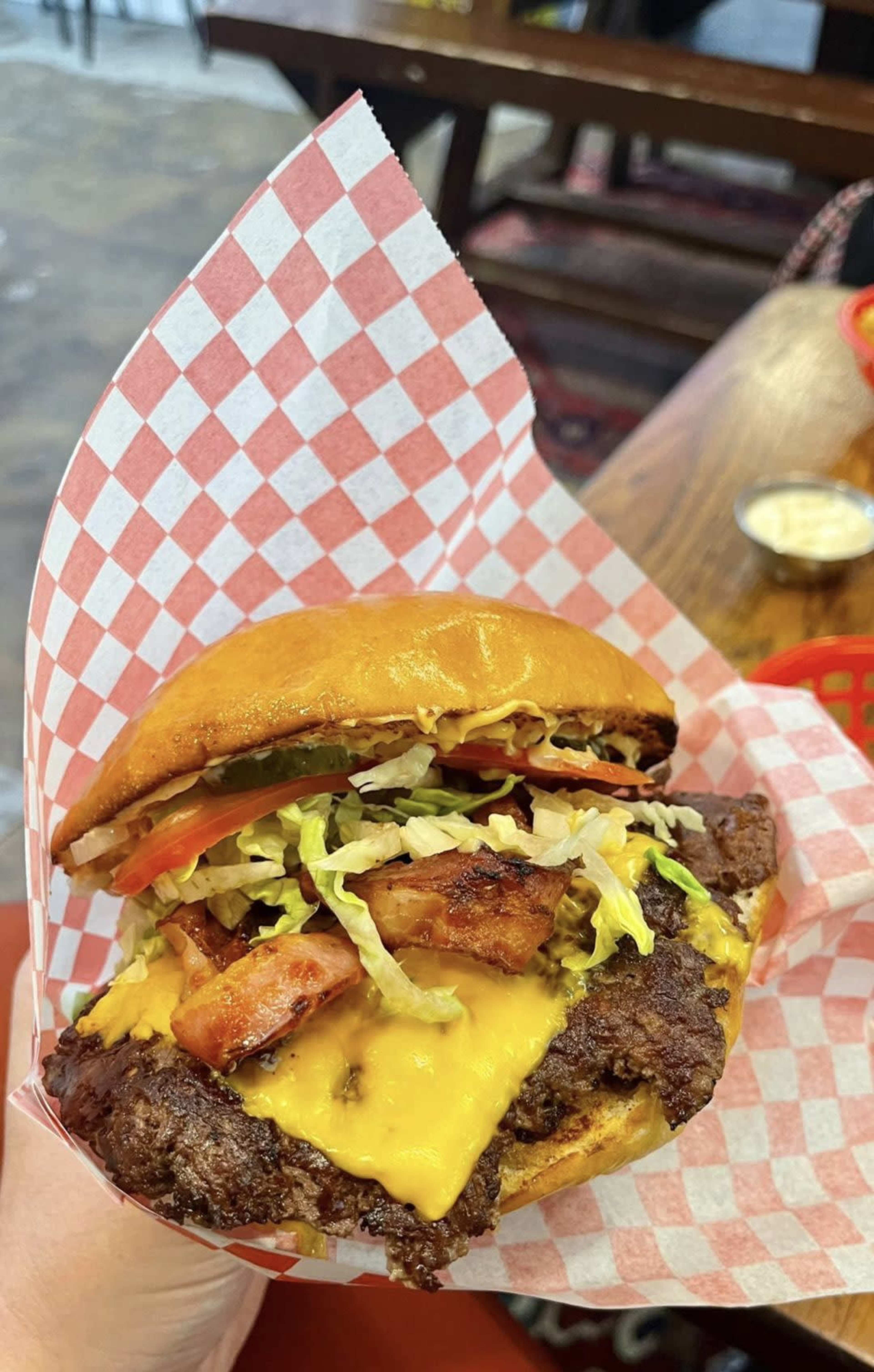 A cheeseburger with lettuce, tomato, pickles, and grilled chicken is held in a hand, placed in a red and white checkered paper liner.