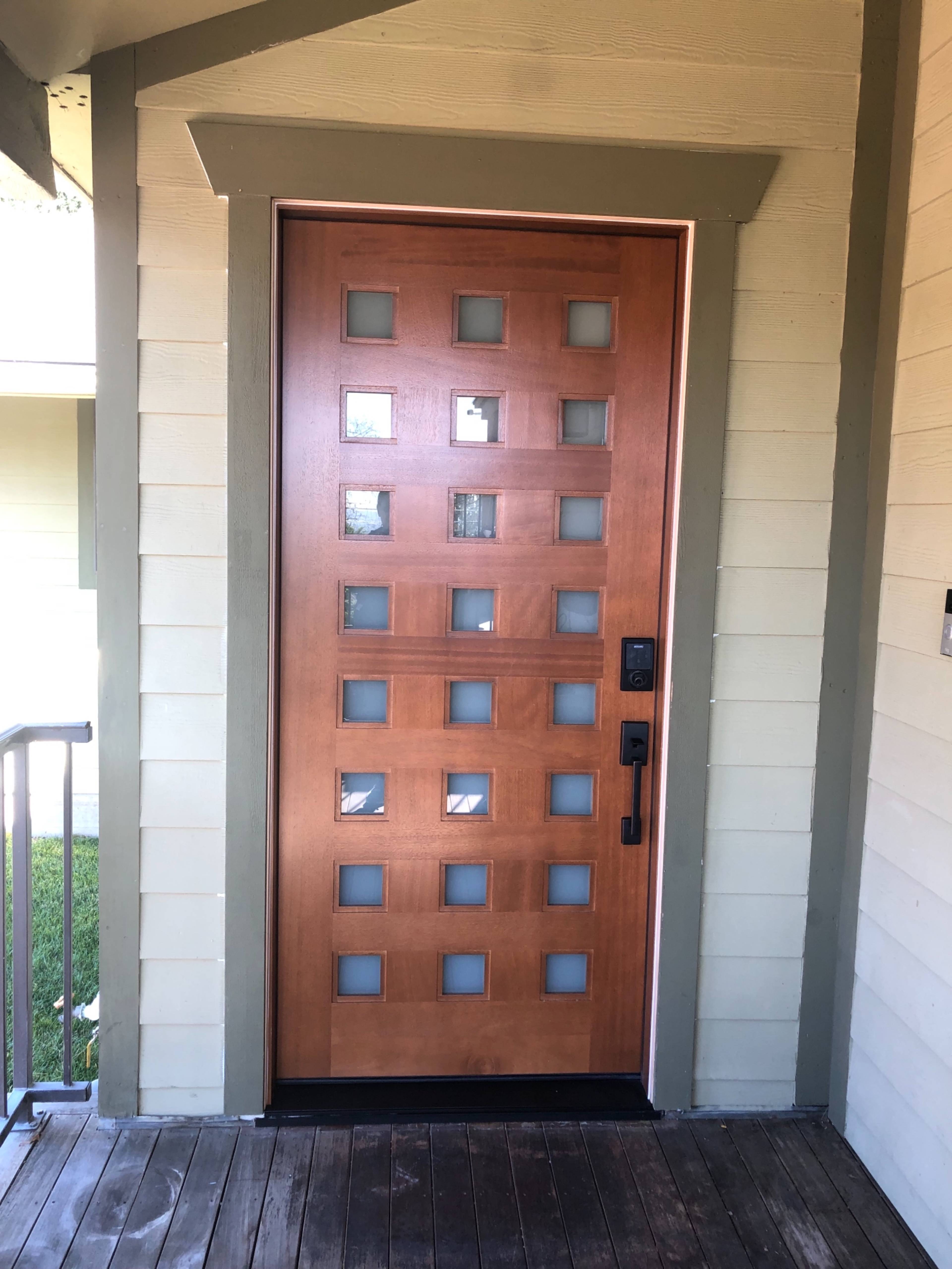 The image shows a wooden front door with square cut-outs and a sleek black door handle, set within a light-colored house exterior.