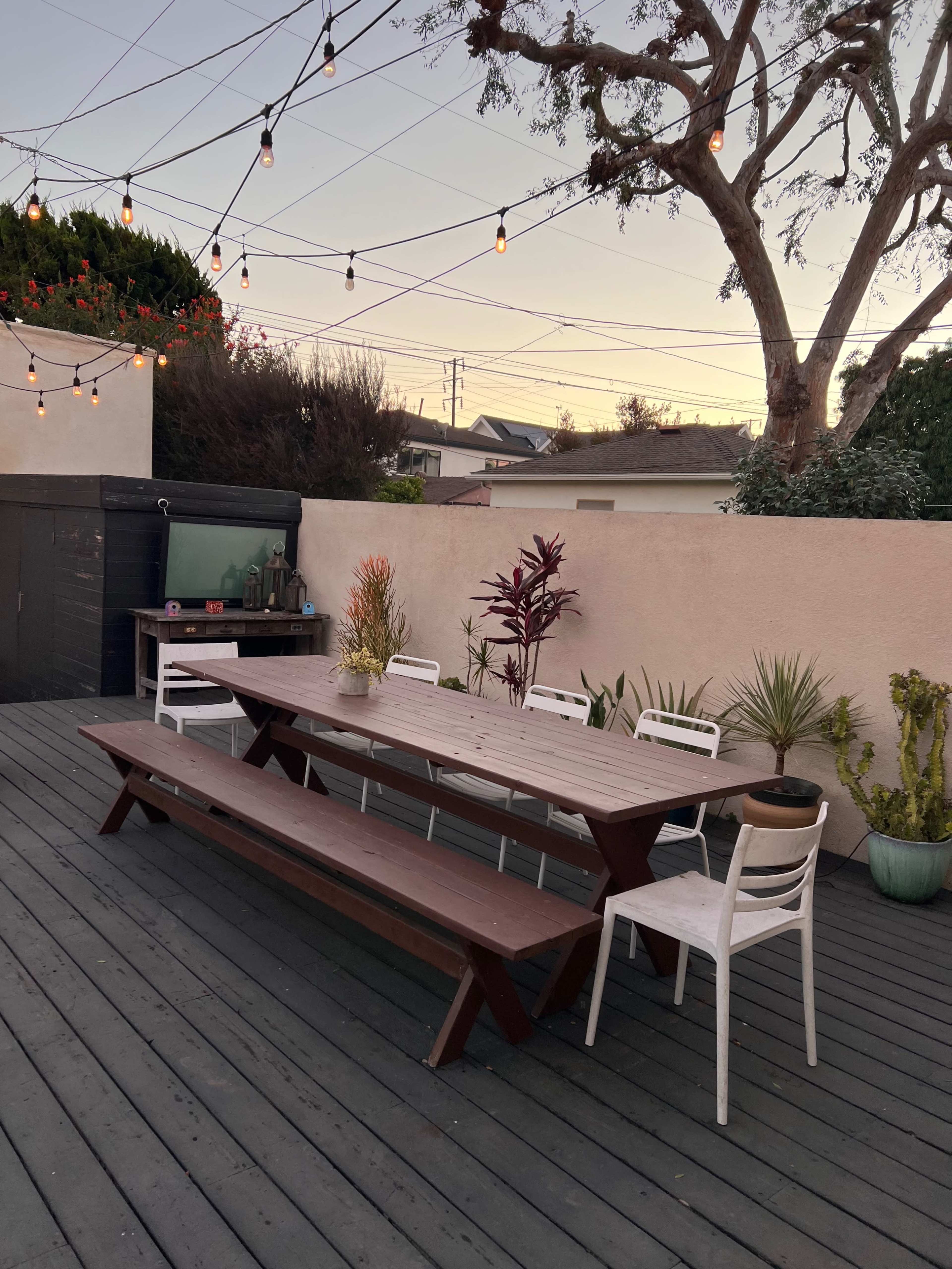 A wooden dining table with benches is set on a deck surrounded by potted plants and illuminated by string lights against a sunset sky.