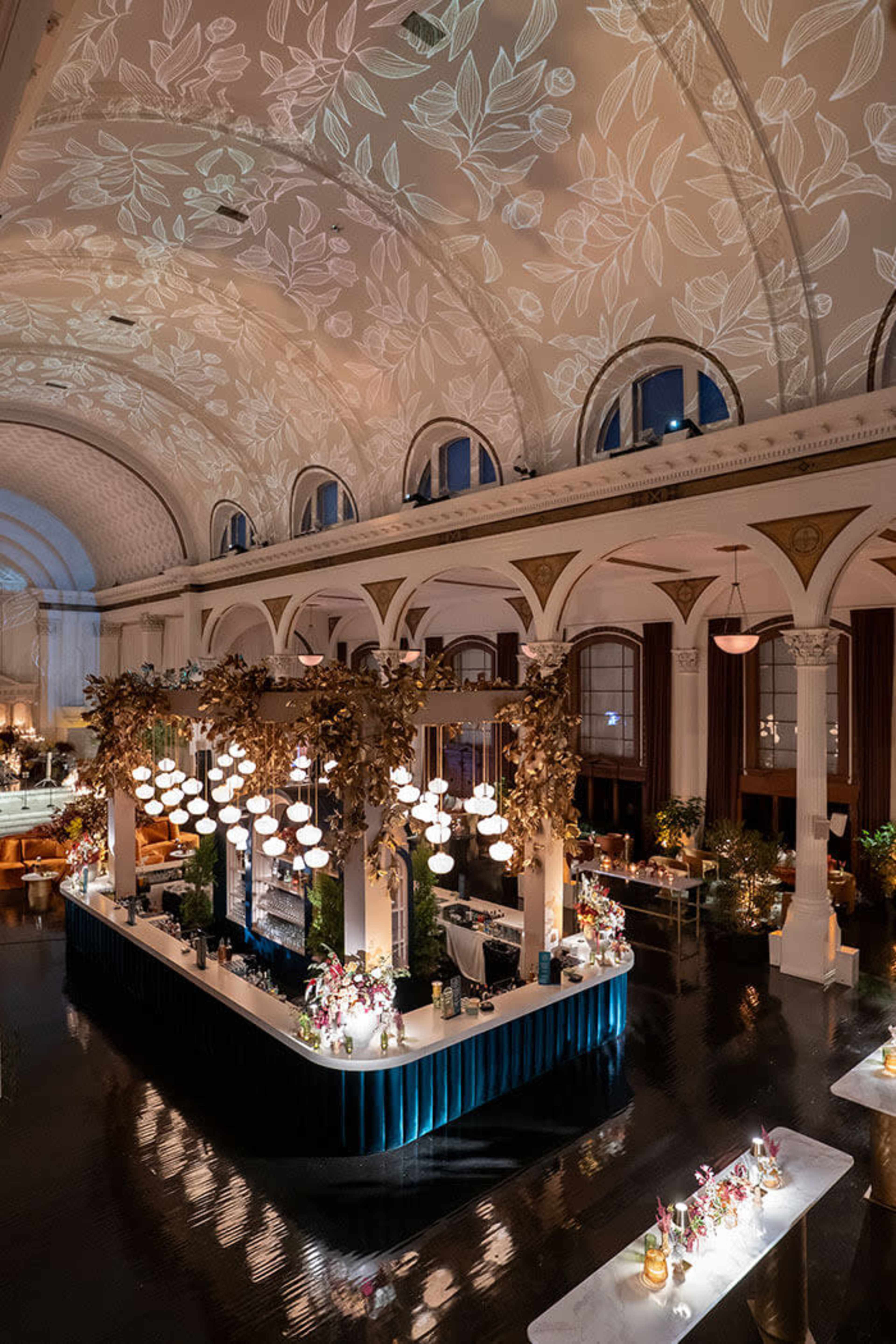 The image shows a luxurious restaurant interior with high ceilings, elegant lighting, and a bar decorated with plants and floral arrangements.